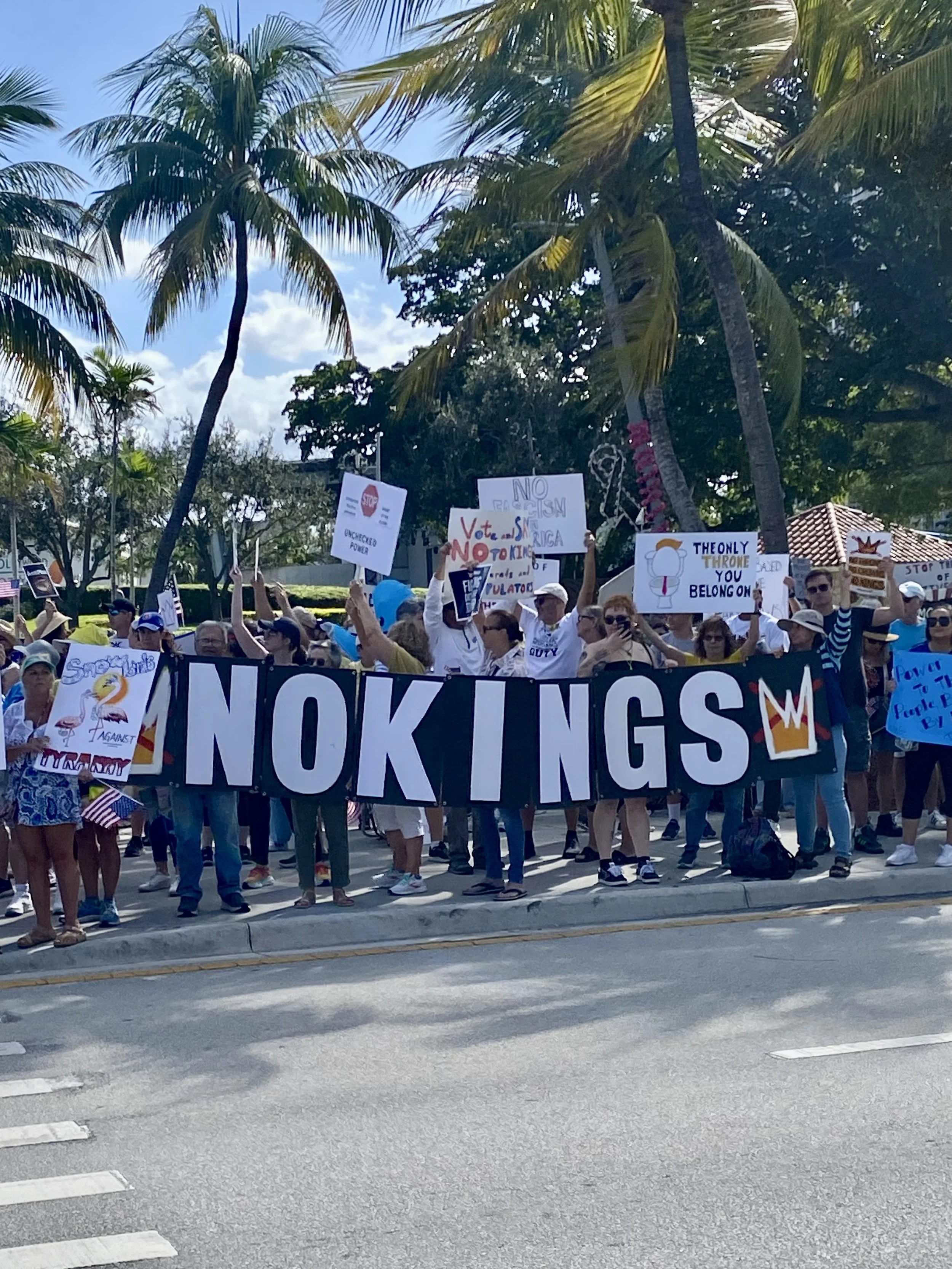 A diverse group of people participating in a protest or rally outdoors, holding various signs and banners. The large banner in the front reads 'NO KINGS.' Some signs include messages like 'Stop' and 'The only throne you belong on.' The event takes place on a sunny day with palm trees in the background.