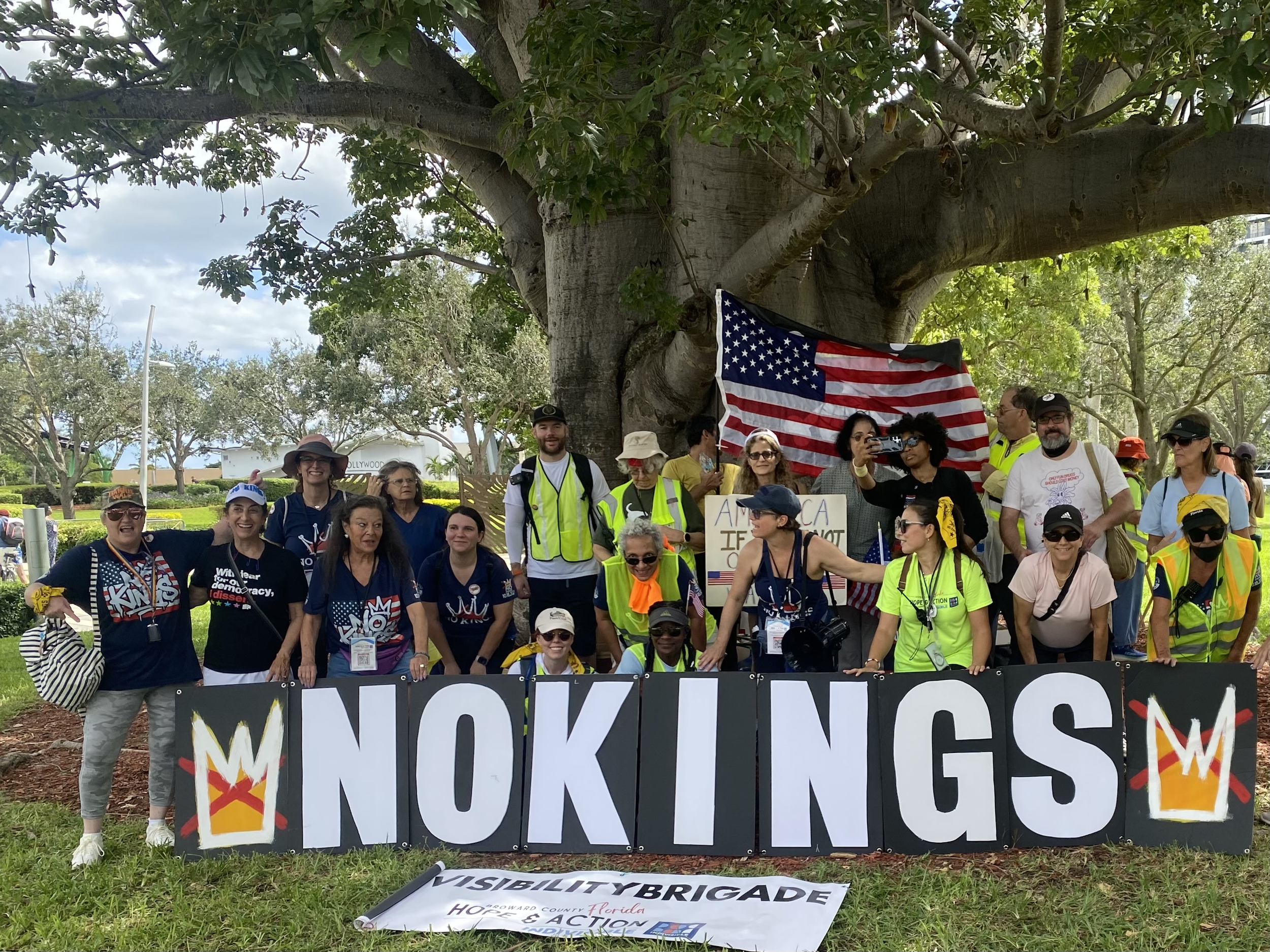 Group of diverse people participating in a protest or rally outdoors, standing in front of a large sign that says "MUNNINGS" and holding an American flag, with others holding signs and wearing safety vests near a large tree.