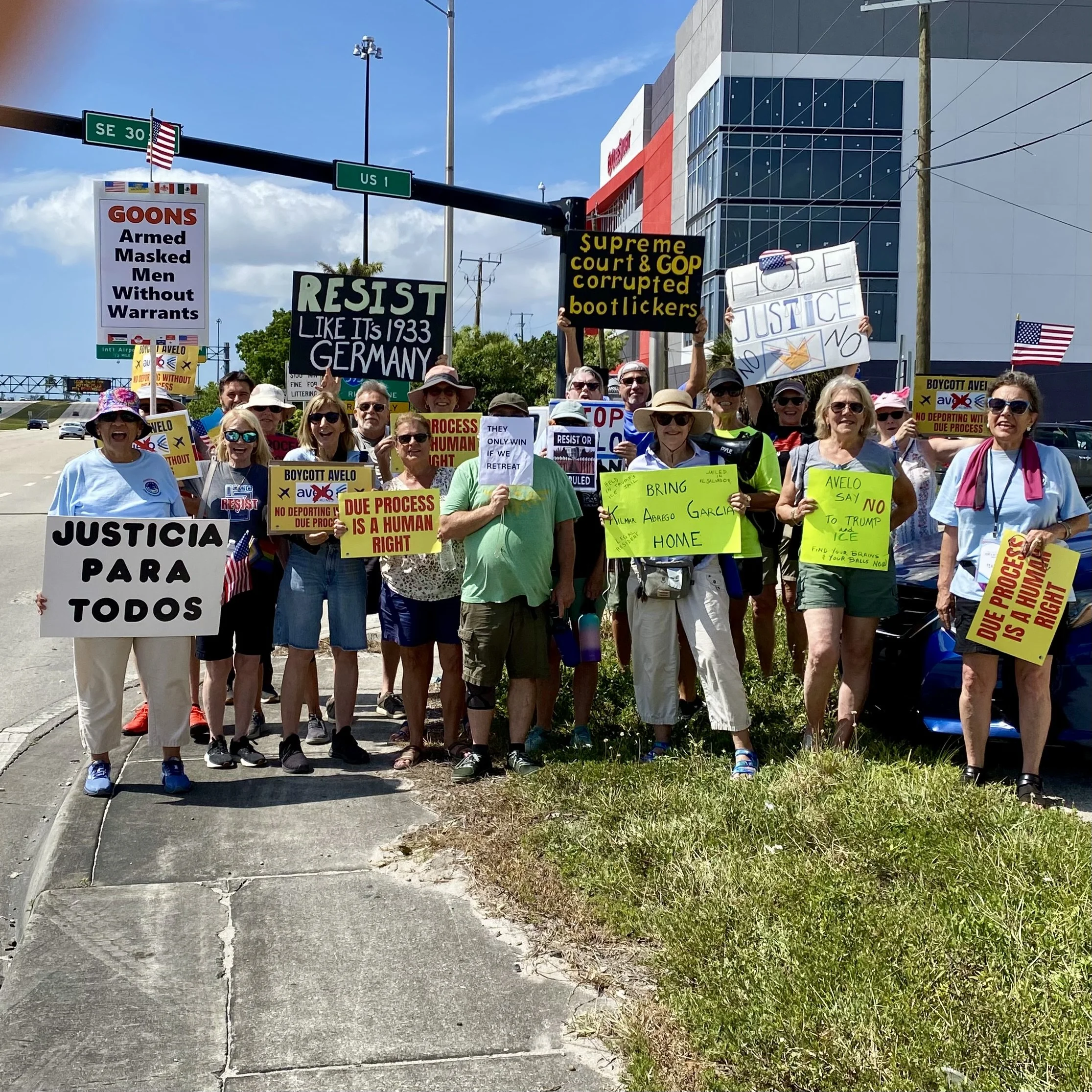 A crowd of people holding various protest signs on a sidewalk, with street signs overhead and a modern building in the background.