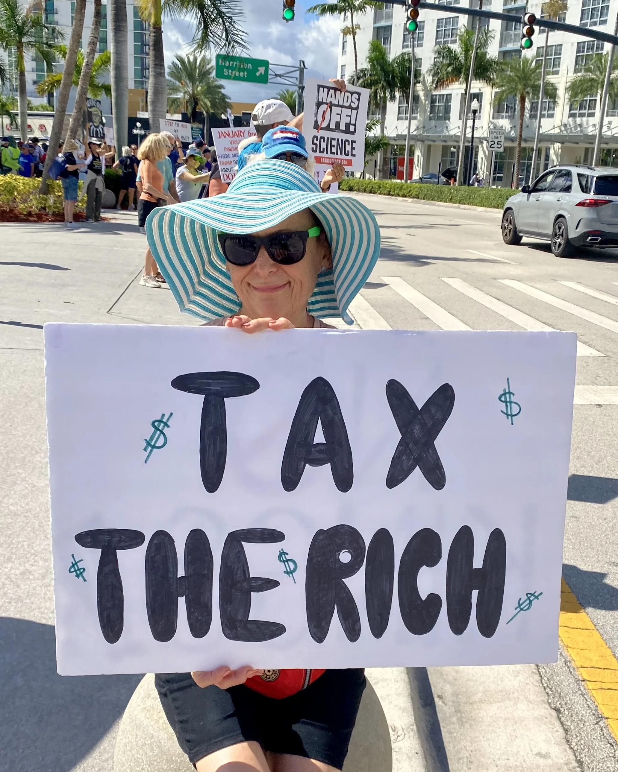A woman at a protest holding a large sign with the words 'TAX THE RICH' written in black letters, and dollar signs surrounding the text. She is wearing a large, wide-brimmed, blue and white striped sun hat and sunglasses, with a background of other protesters, office buildings, and palm trees.