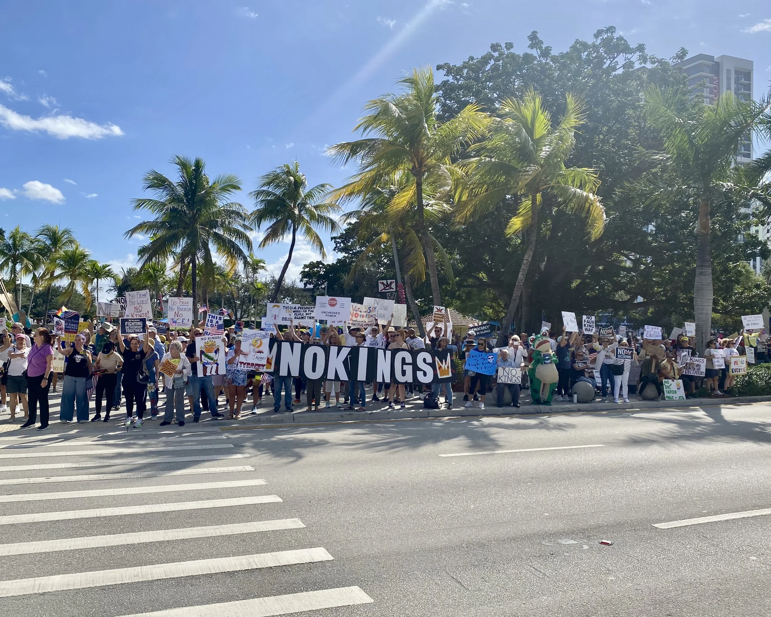A crowd of protesters holding signs and banners during a demonstration under a bright blue sky with palm trees and a tall building in the background.