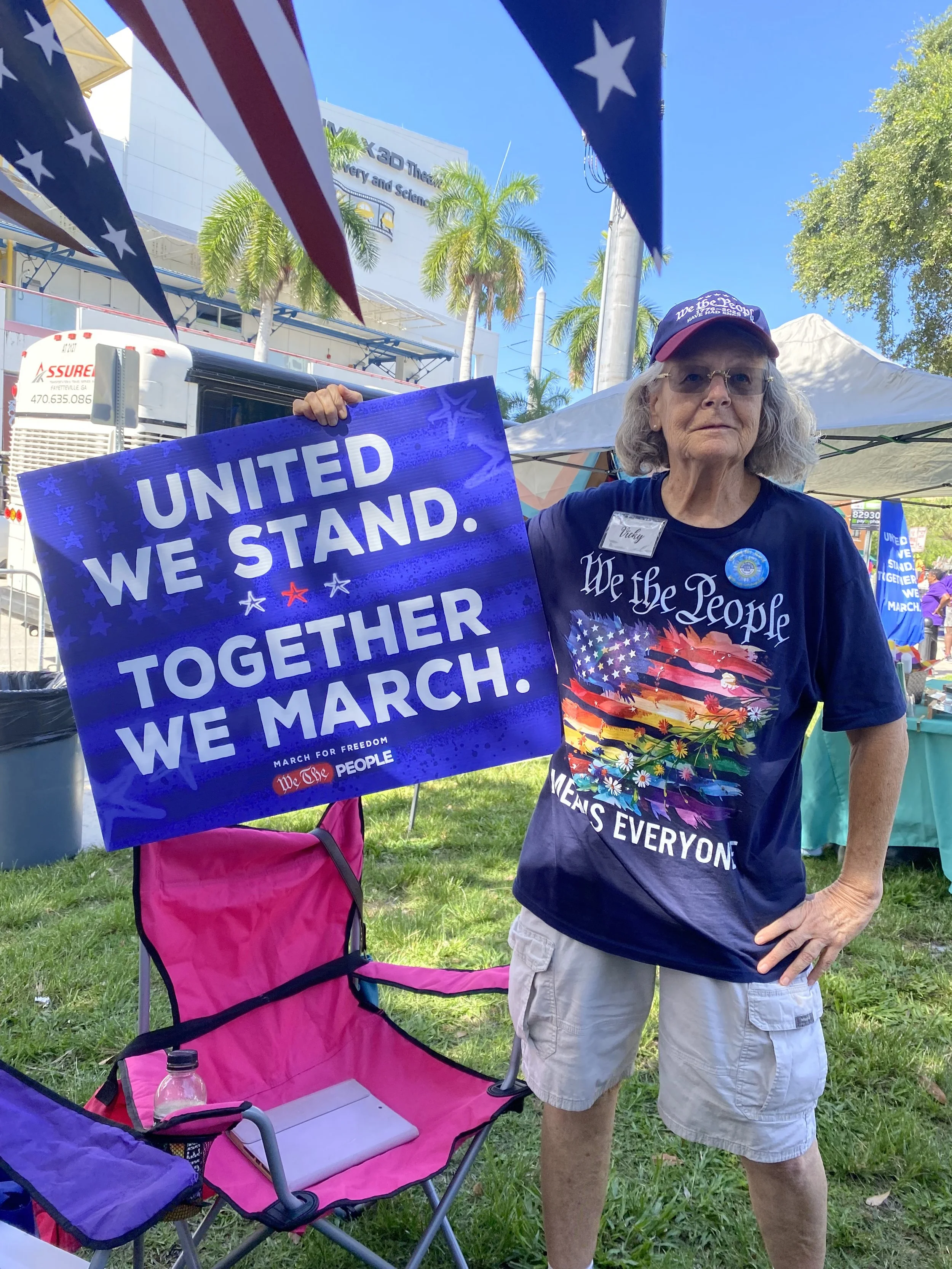 An elderly woman at an outdoor march event holding a sign that says 'United we stand, together we march.' She is wearing a patriotic-themed shirt and a red cap. The background shows tents, a bus, and palm trees under a blue sky.
