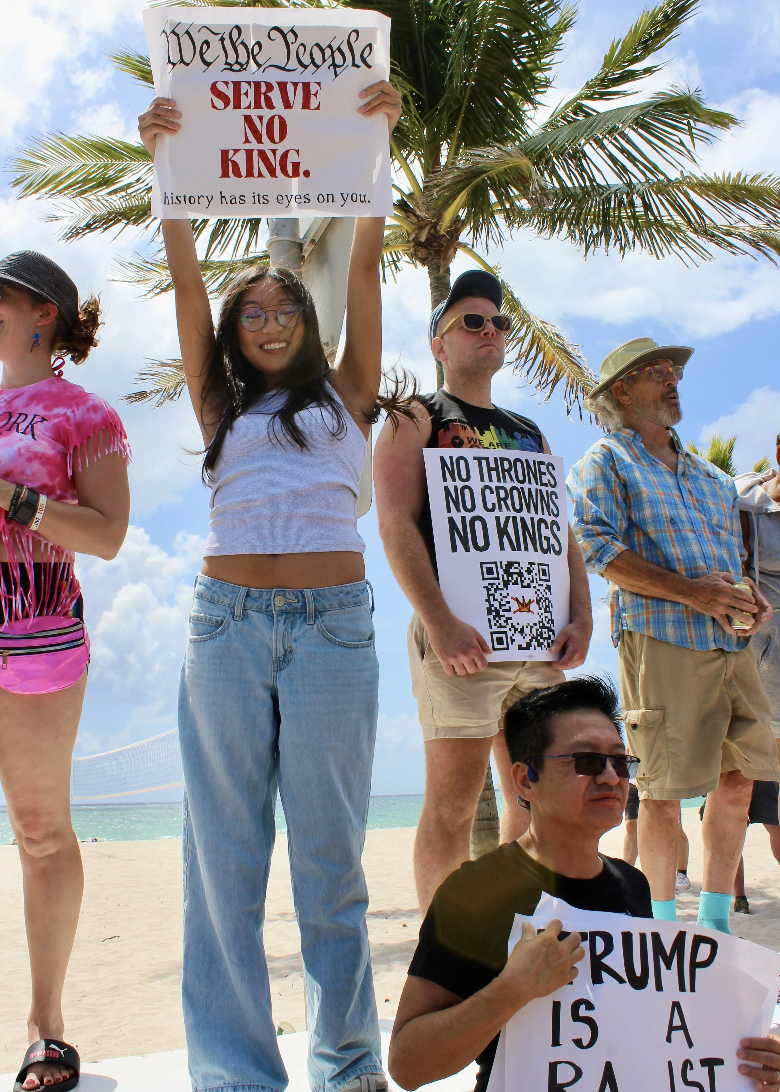 People holding protest signs on a beach, including one that reads, 'We the People serve no king. History has its eyes on you.'