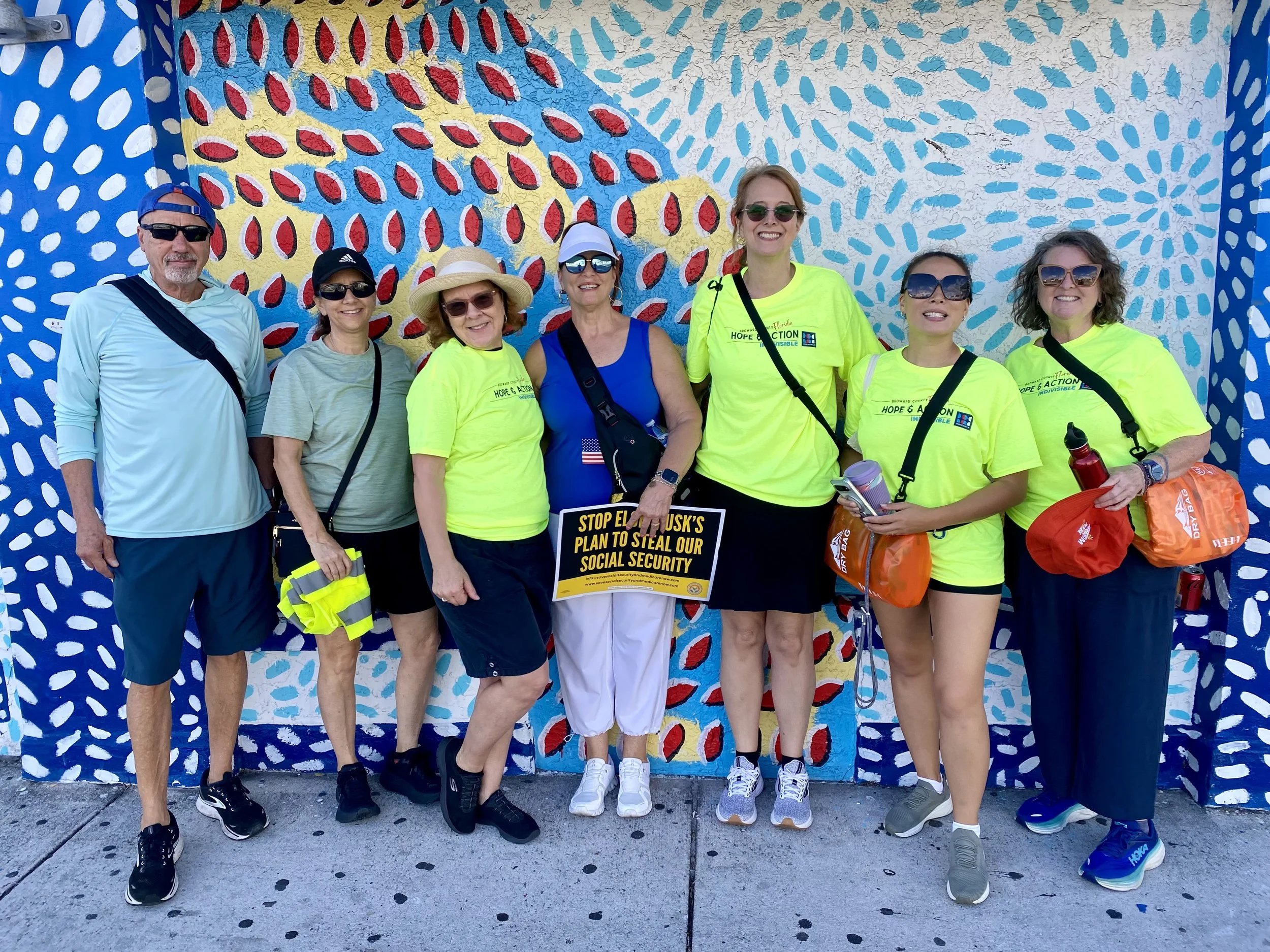 Group of seven people standing in front of a colorful mural, smiling. Four women are wearing bright yellow T-shirts with event logos, one woman in a blue tank top, and a man in a light blue shirt with sunglasses and a black strap. Some are wearing hats and sunglasses, and they are carrying bags and water bottles.