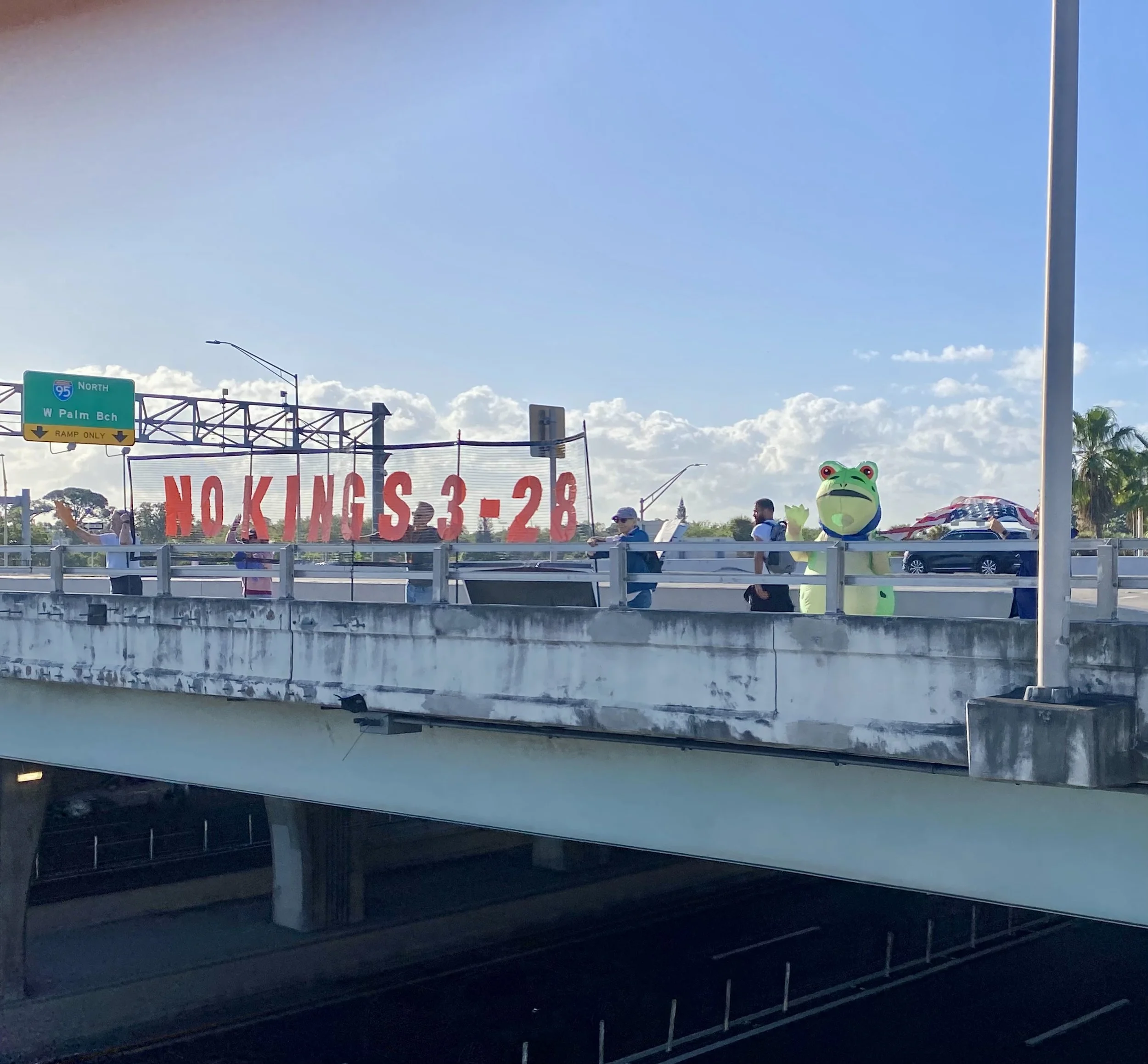 The image shows a portion of a highway with a green overhead sign indicating the northbound lane of I-95 towards W Palm Beach, and a large red and white banner with partially visible text that includes 'NO KINGS 3-28'. There is also a person dressed in a frog costume visible on the right side of the image, along with several other people and cars on the highway.