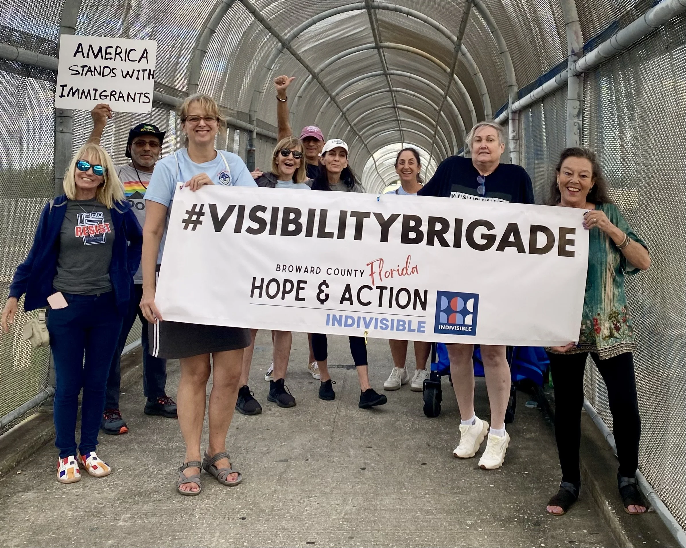 A diverse group of people holding a large banner at a rally or protest, with some women smiling and one woman holding a sign that reads 'America stands with immigrants.' They are standing under a covered walkway with metal fencing on the sides.