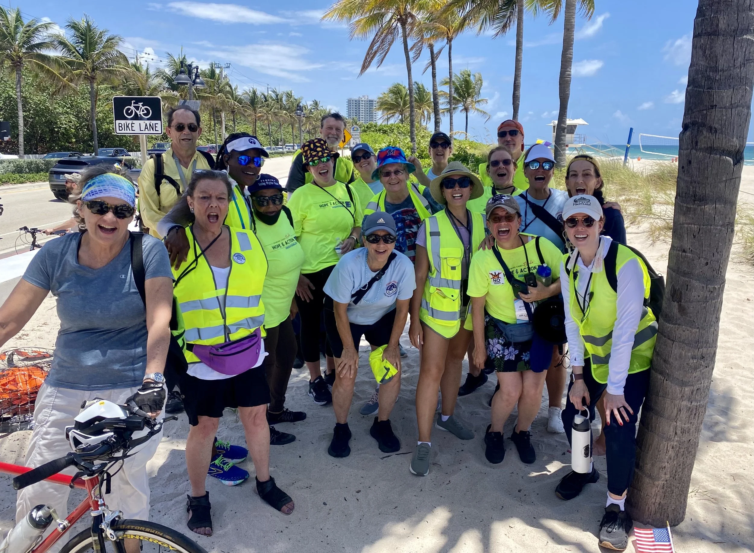A group of smiling people wearing neon yellow safety vests and casual summer clothing gathered together on a beach with palm trees, blue sky, ocean, and volleyball net in the background.