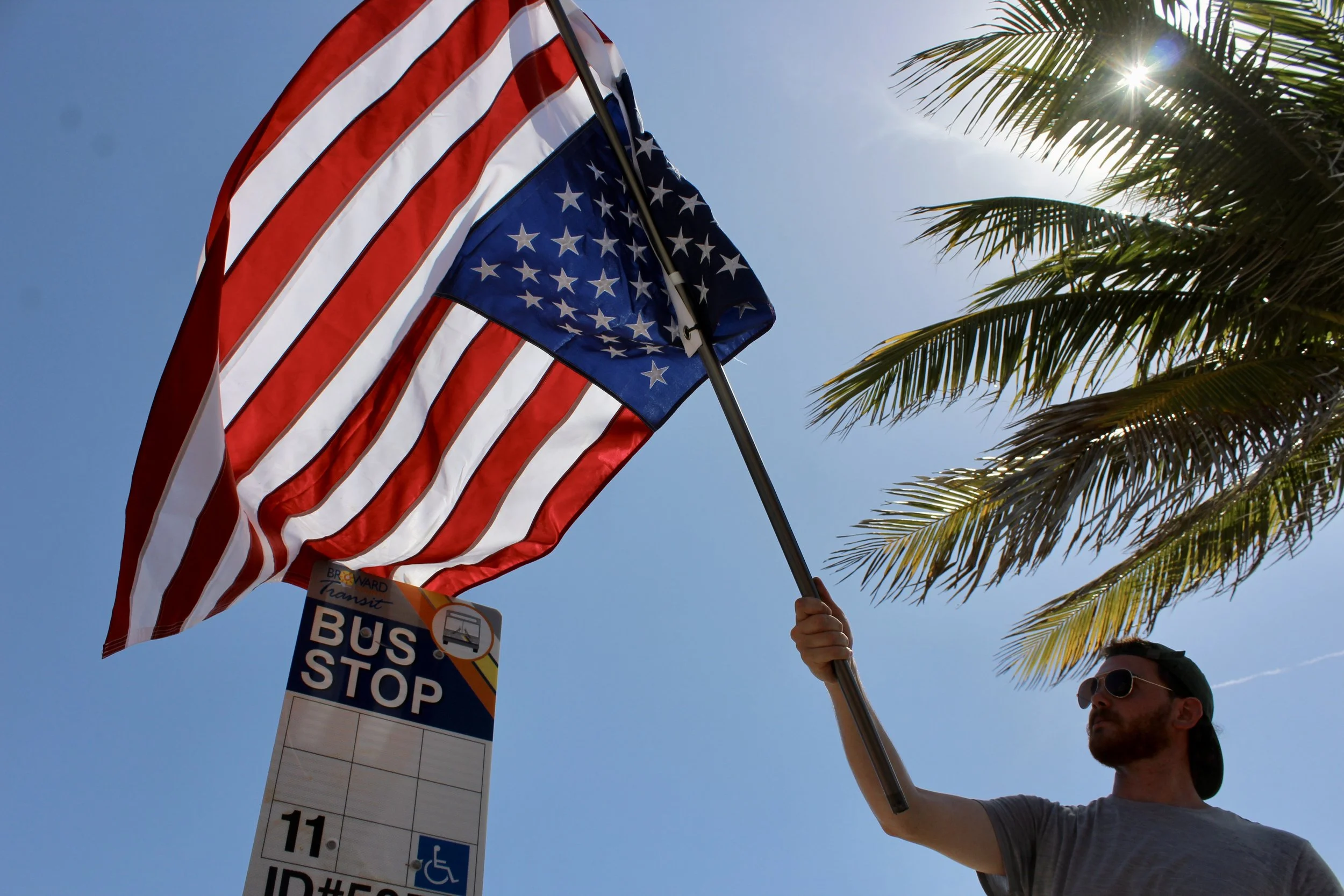 A person holding an American flag on a pole near a palm tree with the sun shining through the fronds, a bus stop sign partly visible below the flag.