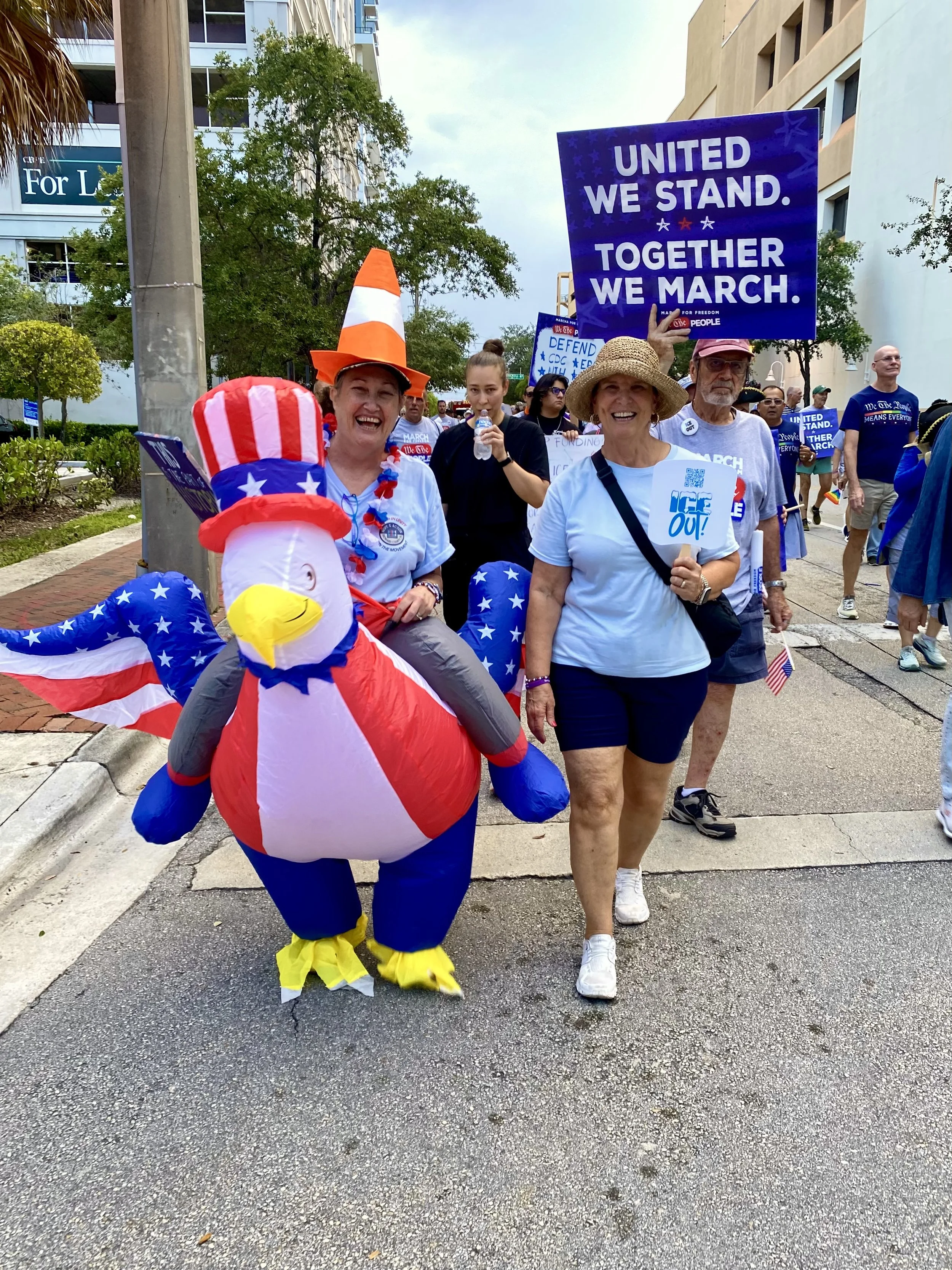 A crowd of people participating in a parade or march, with people holding signs that read 'United We Stand. Together We March.' Some participants wear patriotic clothing and accessories, including a person dressed as an Uncle Sam statue. The scene is outdoors, on a city street with buildings and trees in the background.