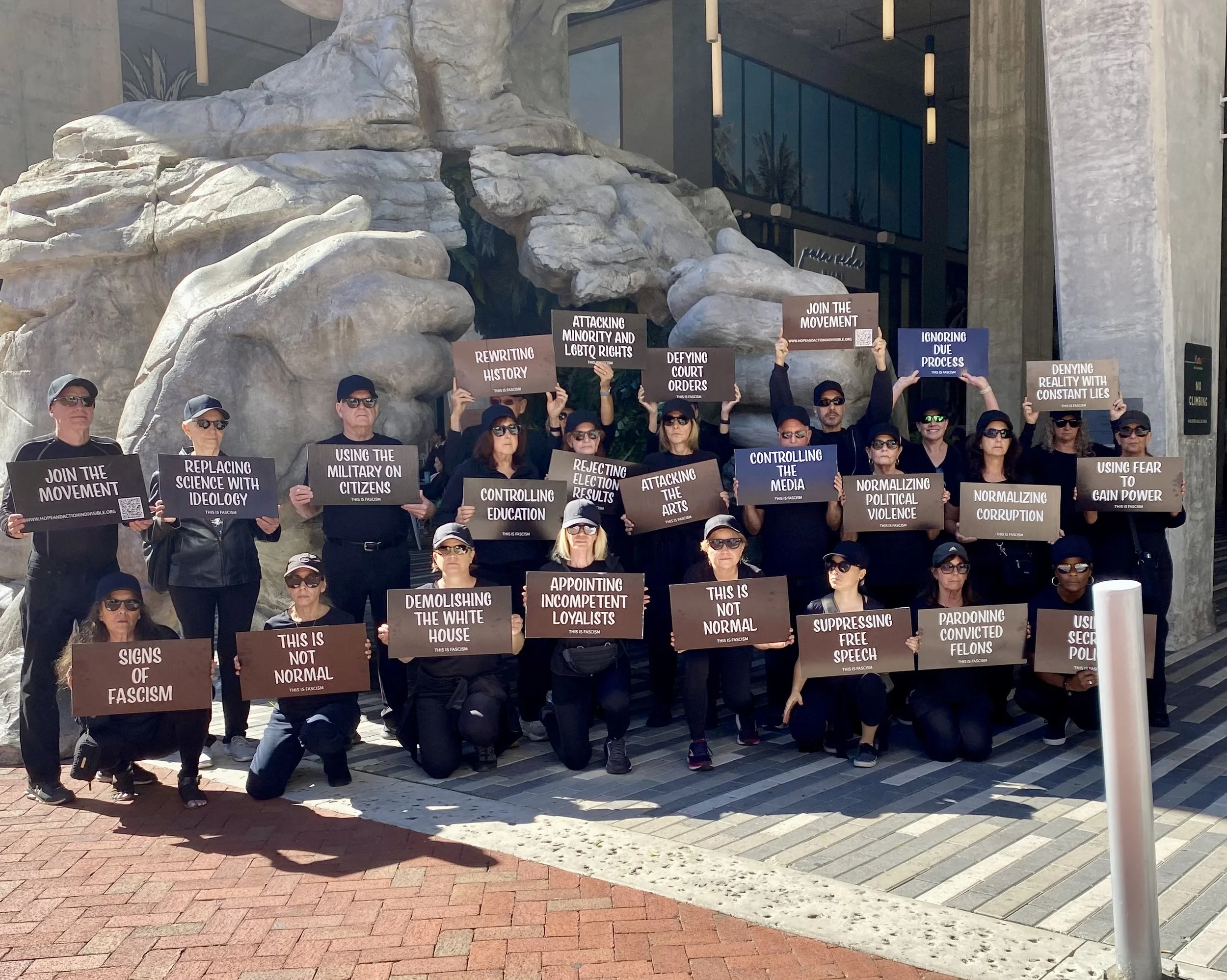A group of protesters holding signs about various social and political issues. The signs include messages such as "Rewriting History," "Attacking Minority and LGBTQ Rights," "Reclassing Science with Ideology," and "Using Fear to Gain Power." The protesters are part of a demonstration against fascism, with some kneeling and others standing, all wearing sunglasses and black clothing, in front of a building with large rocks and modern glass architecture.
