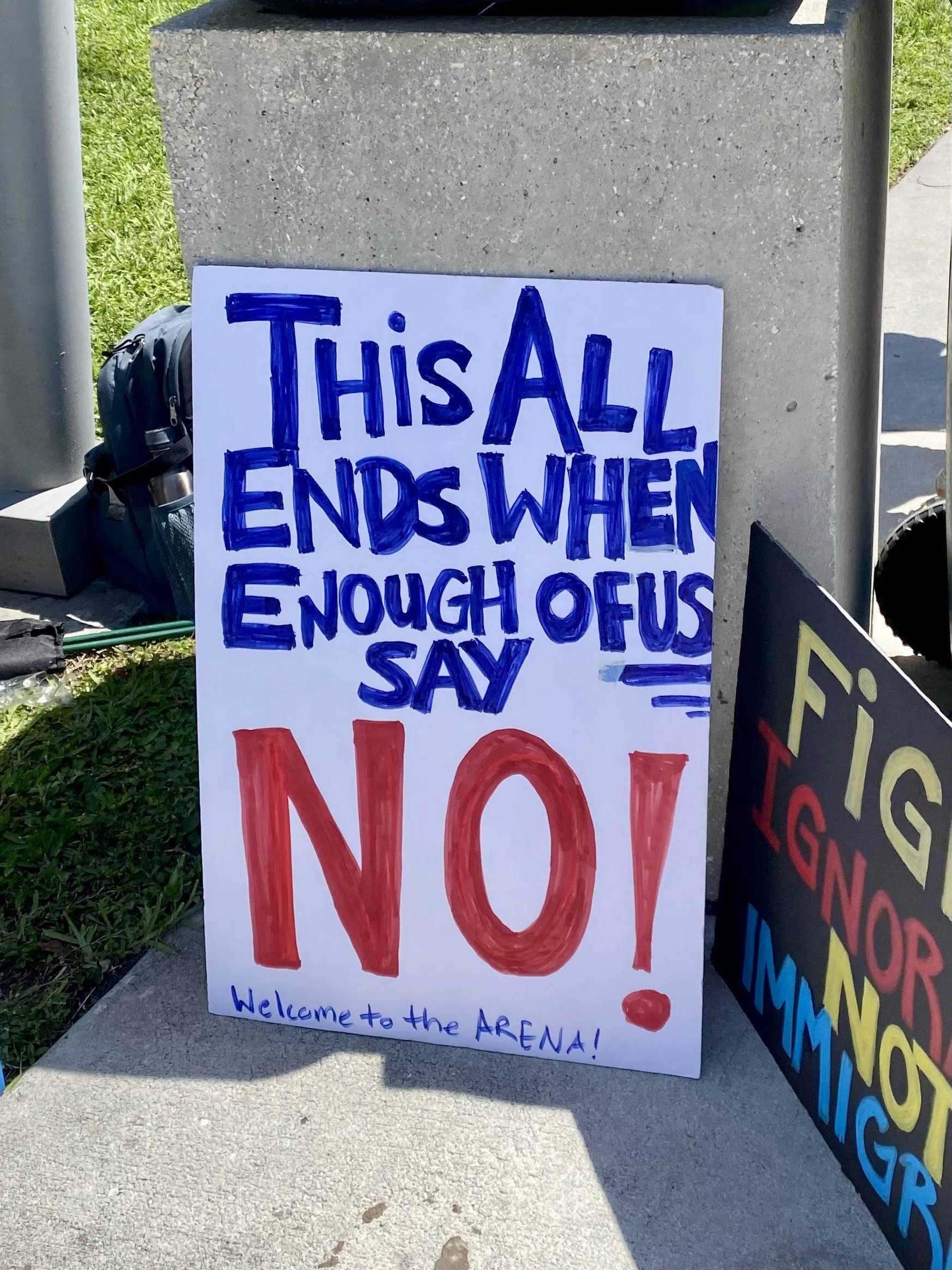 A handwritten sign on a white poster board says, 'This All Ends When Enough of Us Say No!' in bold blue and red letters, with a smaller note at the bottom that reads 'Welcome to the ARENA!' The sign is placed against a concrete surface outdoors, with a backpack and other signs nearby.