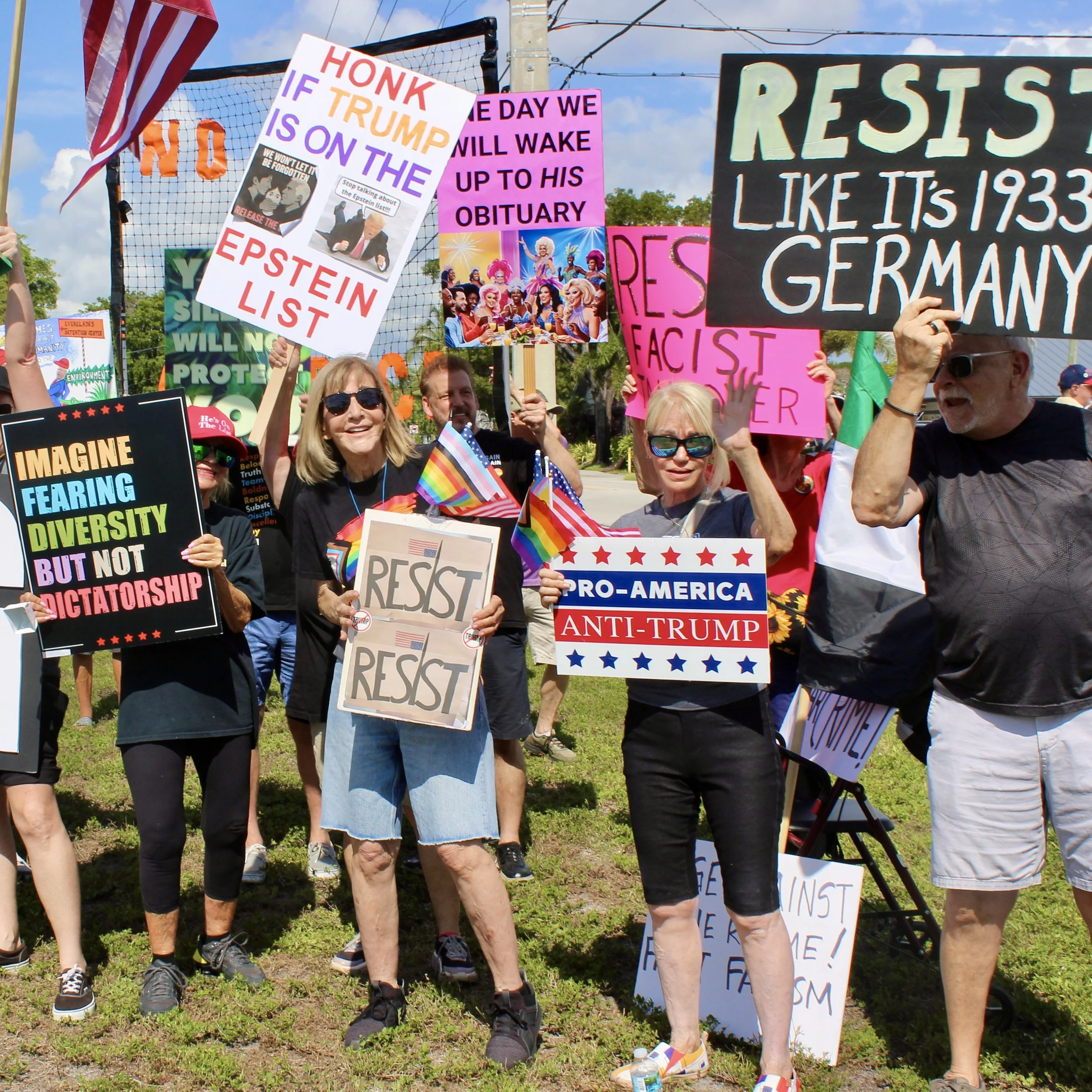 A group of protesters holding signs at a rally. Signs include messages such as 'Honking if Trump is on the Epstein List,' 'We will wake up to his obituary,' and 'Resist, like it's 1973, like Germany.' People are smiling, wearing sunglasses, and some have rainbow flags.
