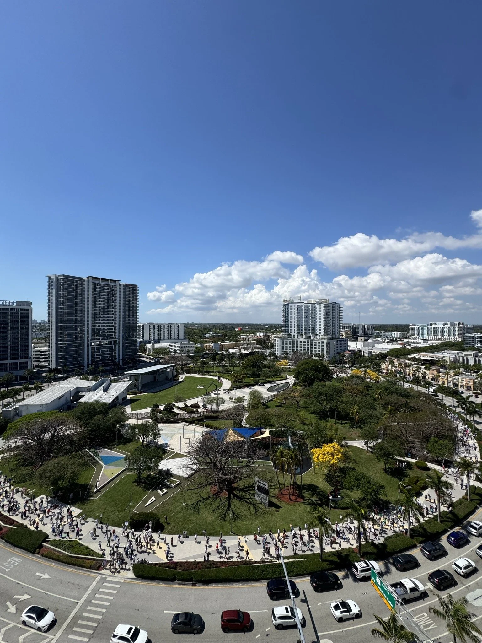 High-angle view of a city park with people walking along pathways, surrounded by tall modern buildings under a bright blue sky with scattered clouds.