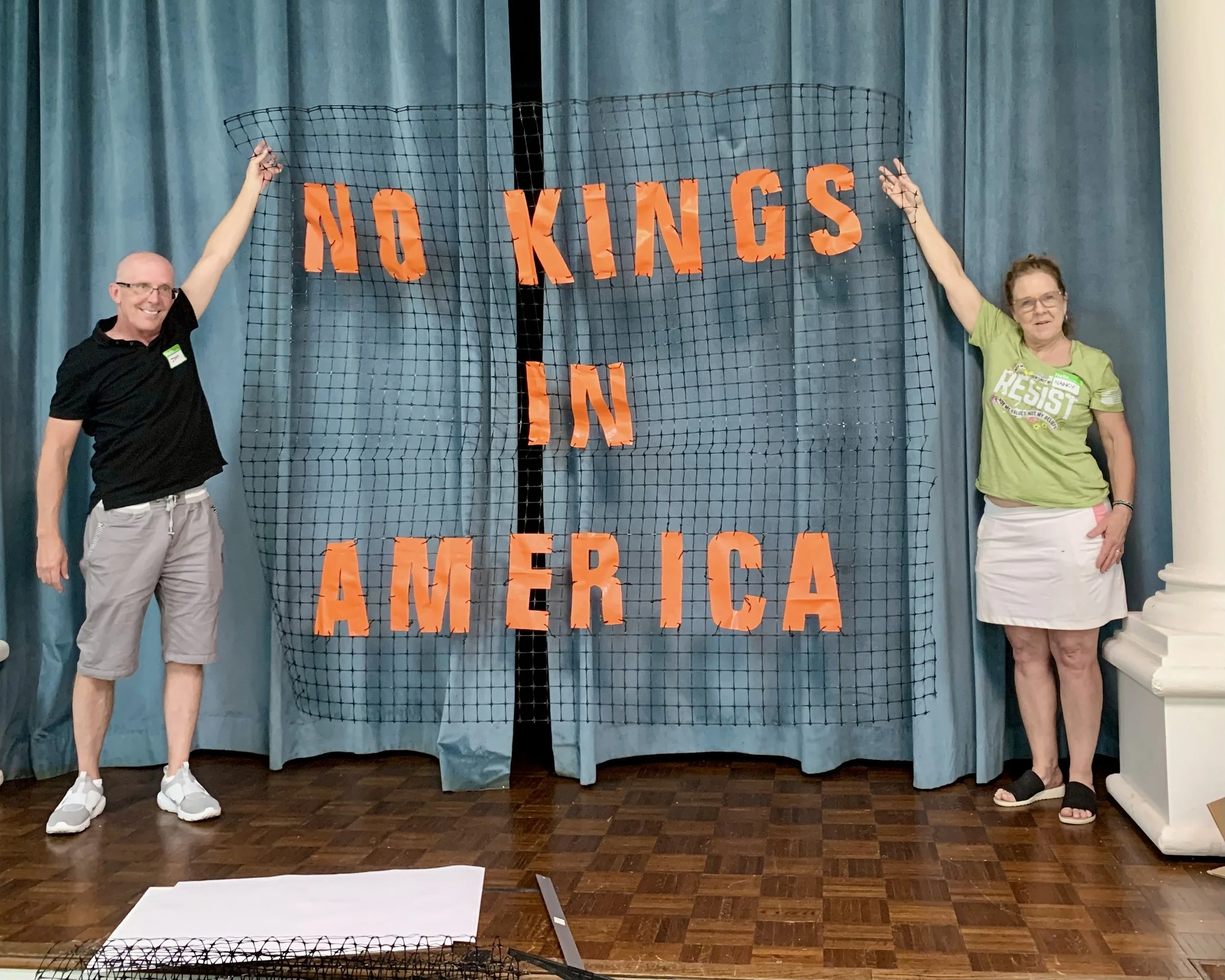 Two people holding a mesh board with the words 'NO KINGS IN AMERICA' written in large orange letters, standing in front of blue curtains.
