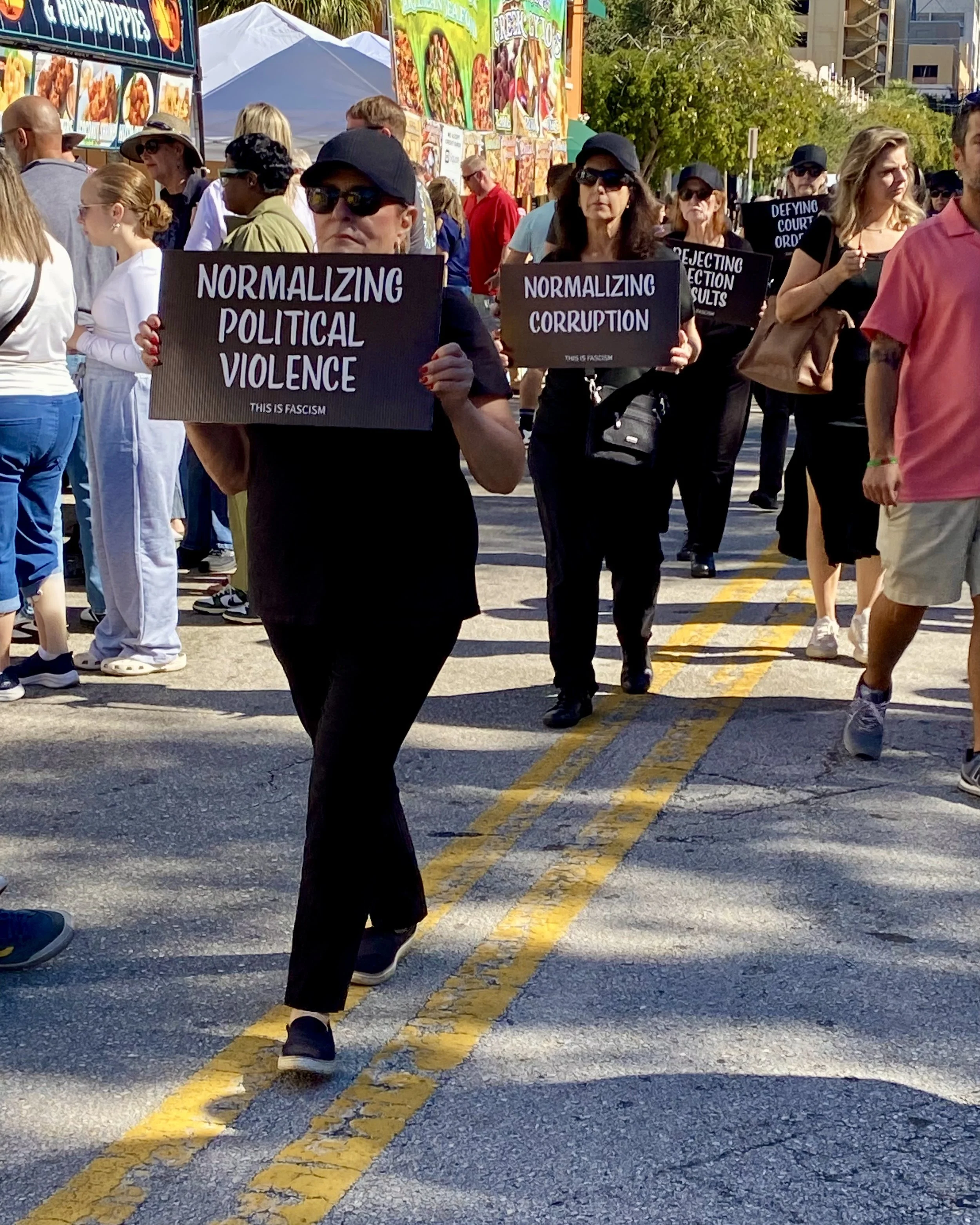 A group of protesters at an outdoor event holding signs that read "Normalizing Political Violence," "Normalizing Corruption," and other messages. People are wearing casual clothing, some with hats and sunglasses, and they are standing on a paved street with a double yellow line. In the background, there are food stalls and trees.