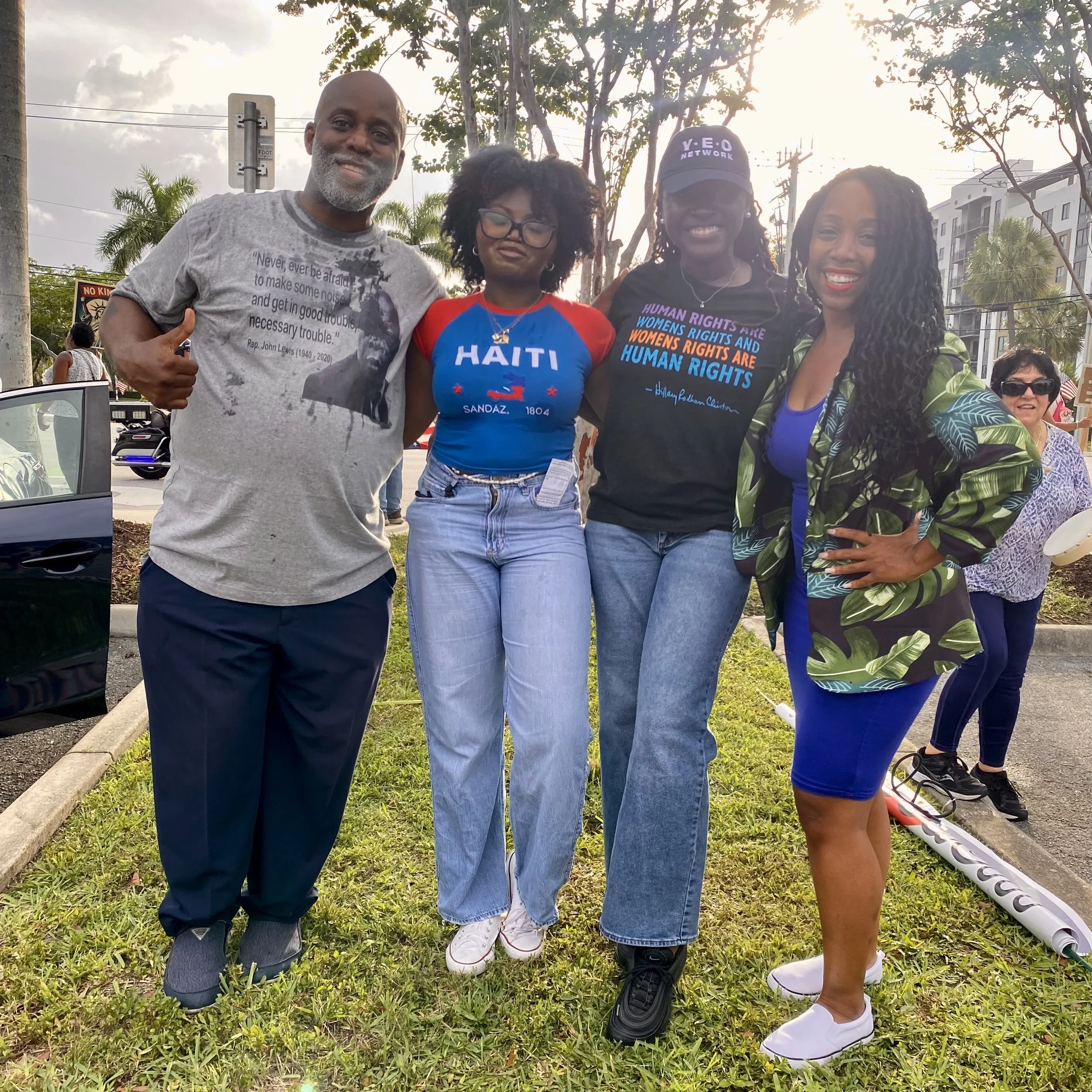 A group of four diverse people standing outdoors in a park or gathering area, smiling at the camera. They are dressed casually and appear to be attending a community or social event. The background includes trees, buildings, and power lines, with an overcast sky.