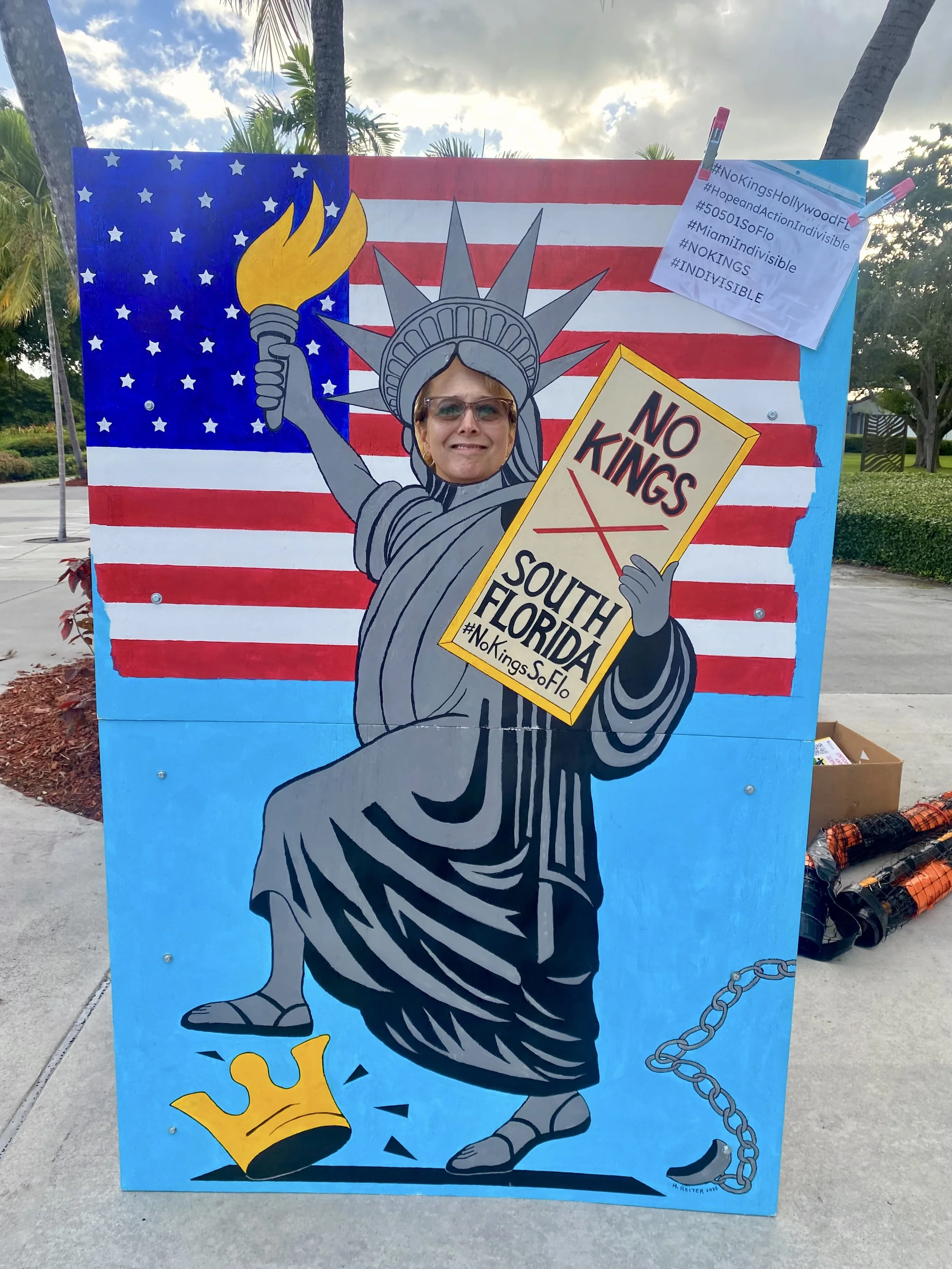 Painting of the Statue of Liberty holding a sign that says 'No Kings South Florida'; background features the American flag; a person with their face visible through the face cutout of the statue in front of the painting.