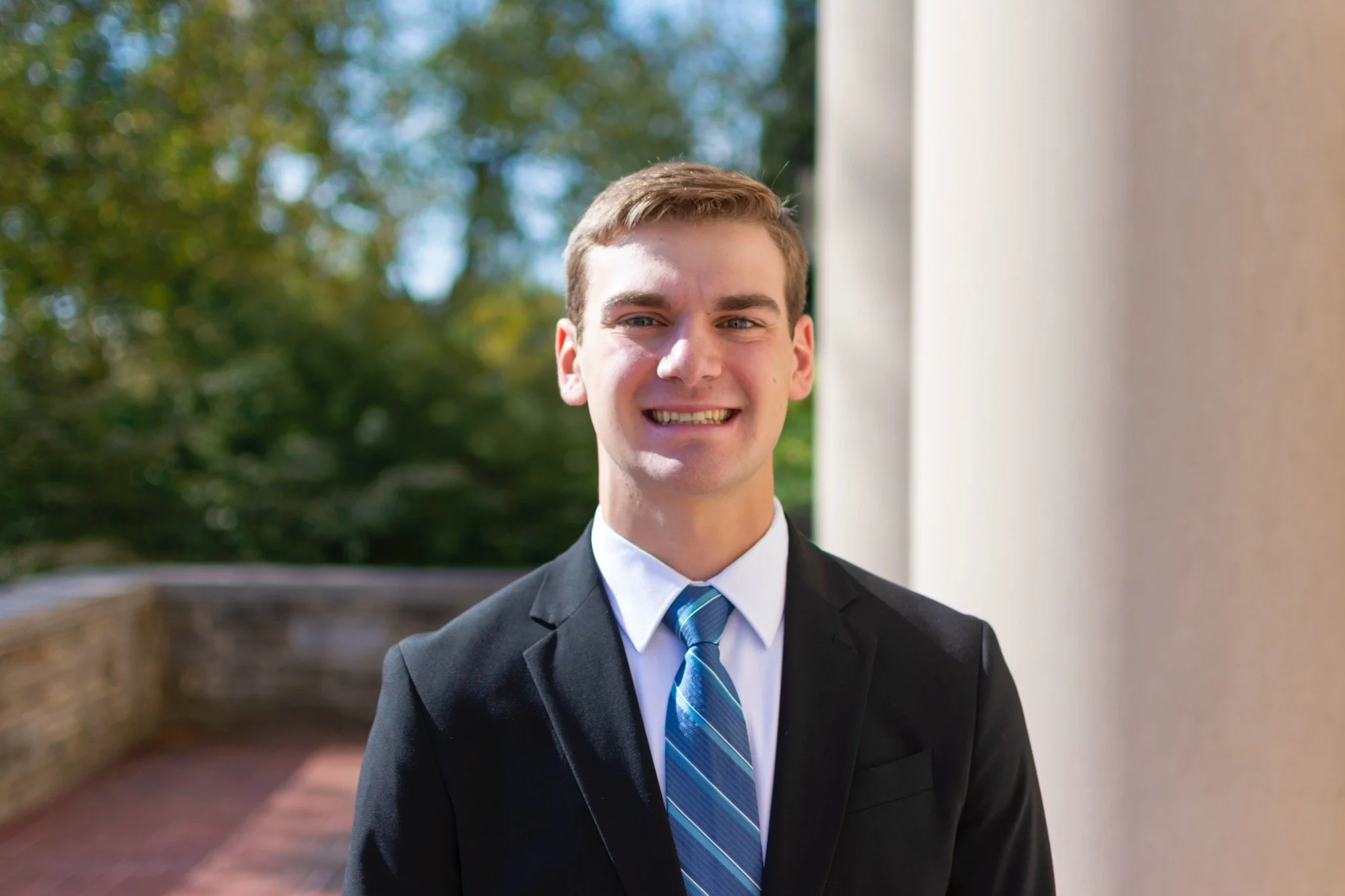 A young man dressed in a black suit with a blue striped tie, smiling outdoors on a sunny day.