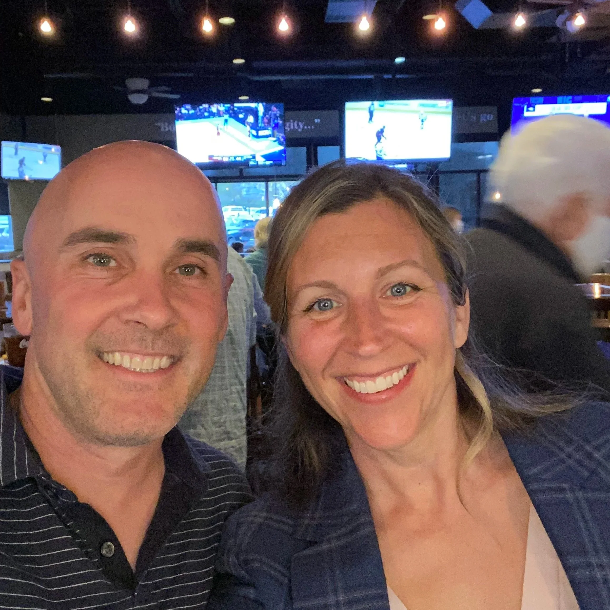 A close-up of a man and a woman smiling, with multiple screens showing hockey games in the background, in a dimly lit sports bar or restaurant.
