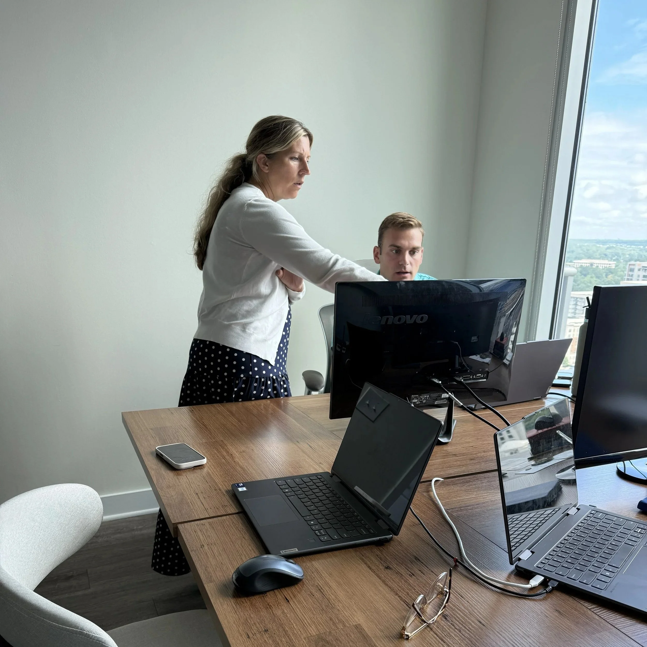 Office workspace with multiple computer monitors, laptops, a smartphone, a mouse, eyeglasses, and a wooden desk. A woman and a man are working together, with the woman pointing at the monitor.