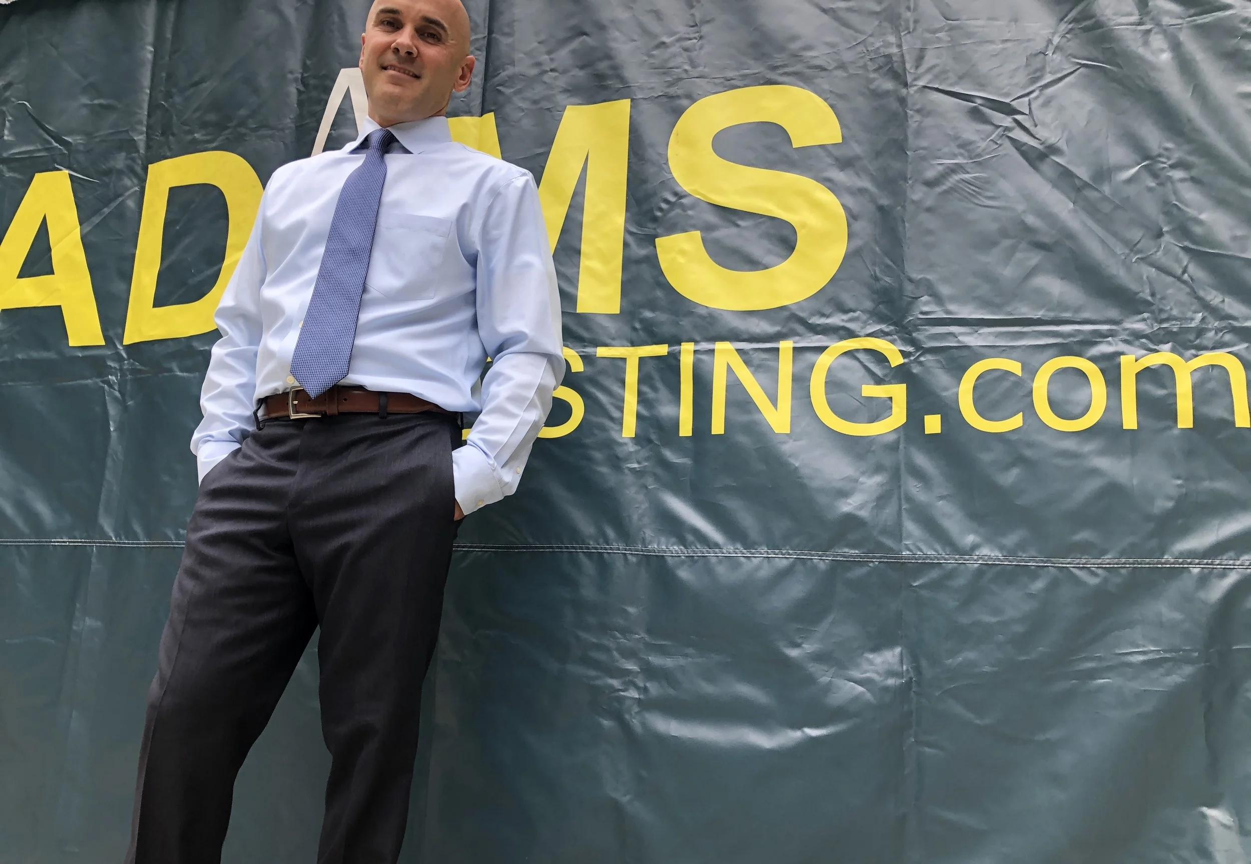 A man wearing a white shirt, tie, and black pants, standing in front of a large banner with yellow and gray text. The banner says 'ADAMSINVESTING.com'.