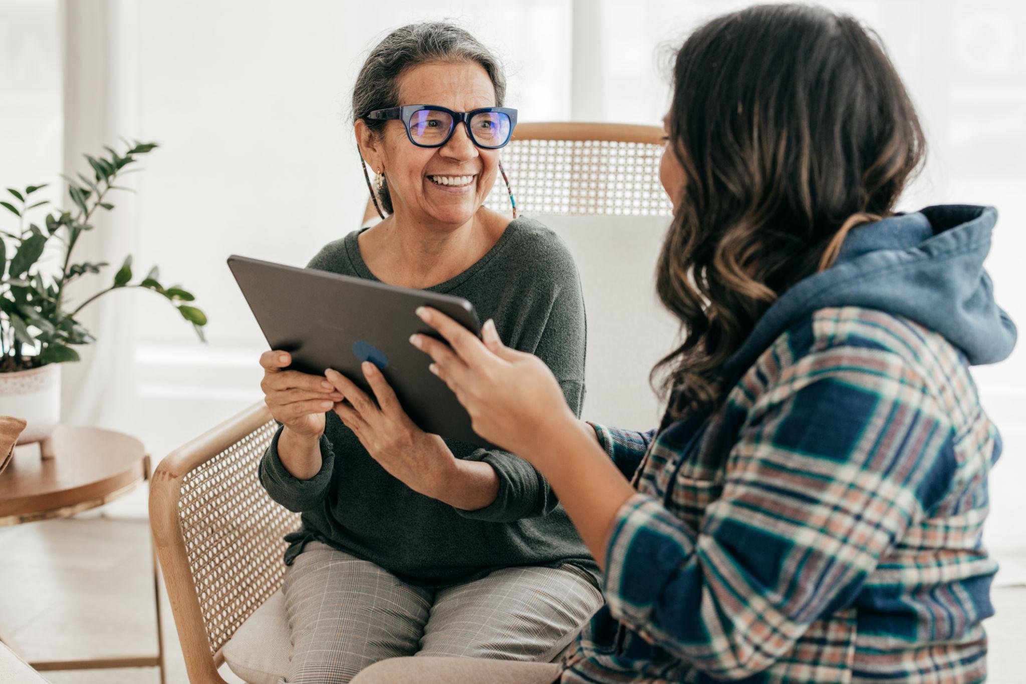 Two women are sitting on chairs, engaging in a conversation. One woman, with gray hair and glasses, is smiling, holding a tablet, and wearing a gray sweater. The other woman, with dark hair, wearing a plaid shirt and hoodie, is facing the first woman. The setting appears to be a bright, modern room with a plant on a side table.