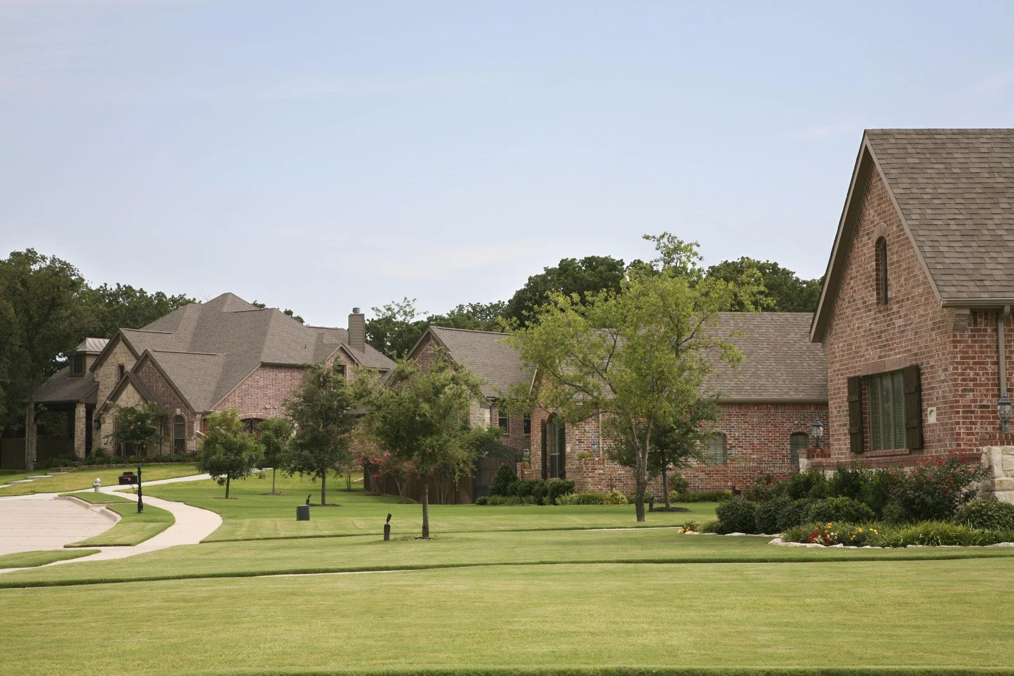 A suburban neighborhood with brick houses, well-maintained lawns, pathways, trees, and a clear blue sky.