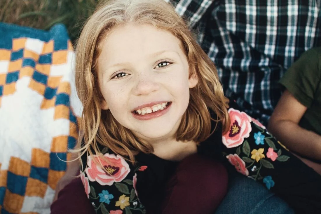 A young girl with red hair, freckles, and a big smile, sitting outdoors near a colorful quilt or blanket.