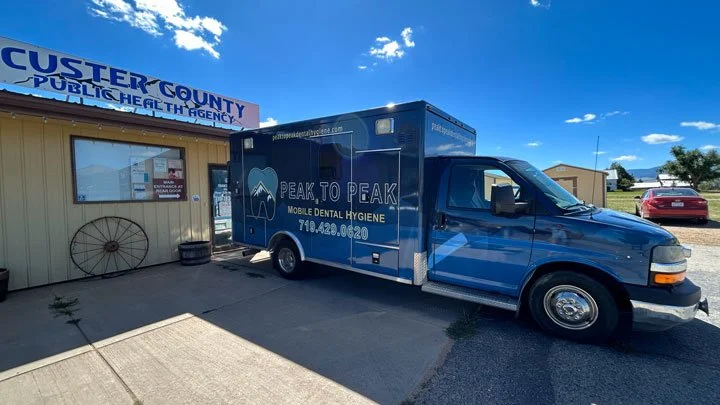 A blue mobile dental hygiene van parked in front of the Custer County Public Health Agency building on a sunny day.