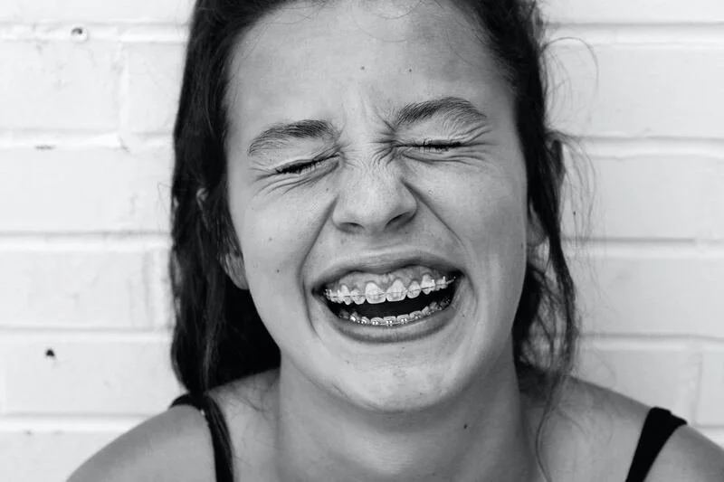 A young woman with braces on her teeth crying with her eyes closed, against a brick wall background.
