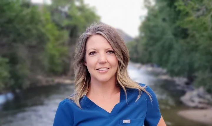 STACI COBERLY Founder of Peak to Peak Dental Hygiene, PC, A woman with shoulder-length blonde hair smiling outdoors near a river with trees in the background, wearing a blue medical scrub top.