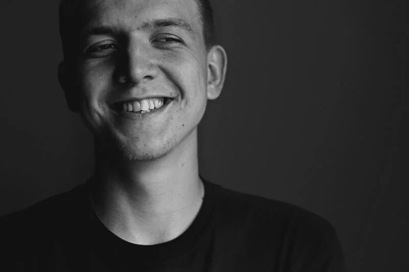 Black and white portrait of a young man smiling, wearing a black shirt, against a dark background.