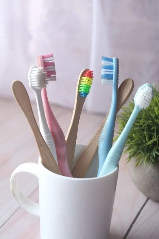 Several colorful toothbrushes in a white mug, with a wooden spoon and a plant in the background.
