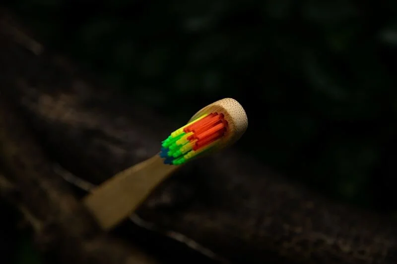 A wooden matchstick with colorful plastic head, featuring red, orange, yellow, green, blue, and black matches, resting on a rough, dark wooden surface.