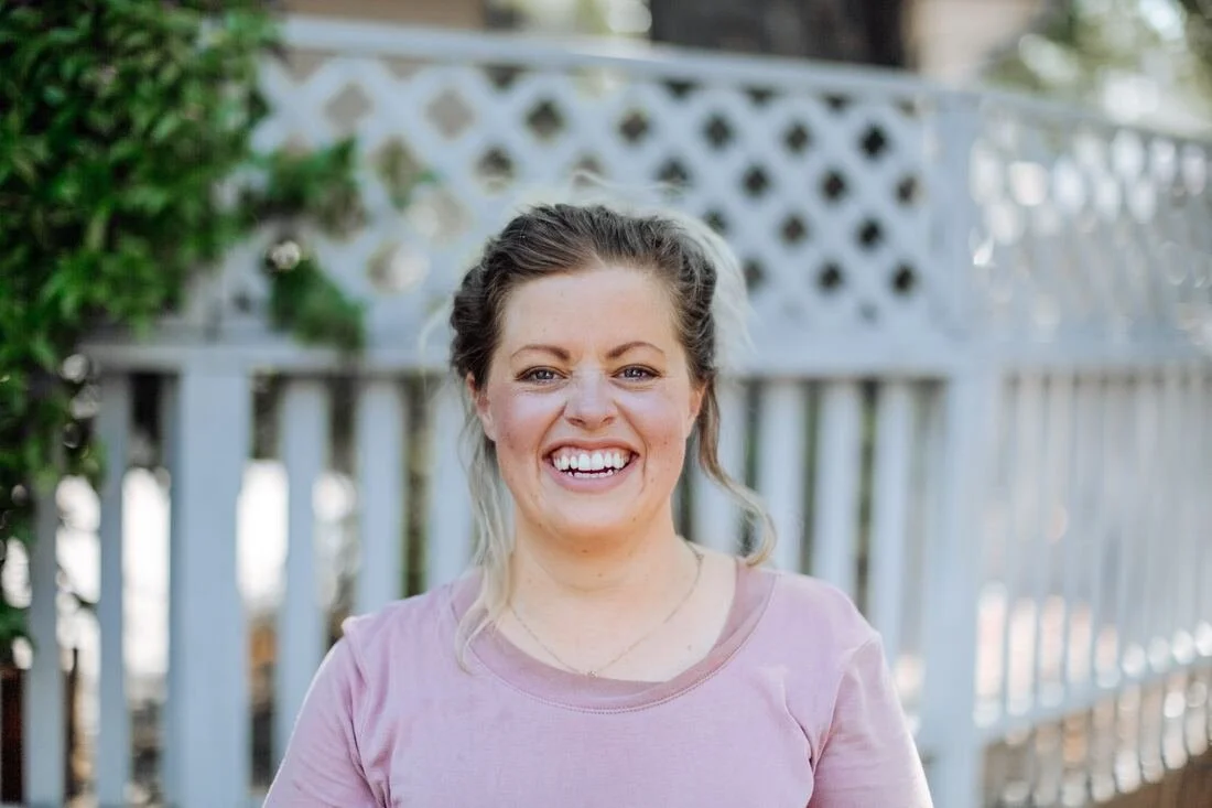 A woman smiling outdoors on a sunny day, with a white fence and greenery in the background.