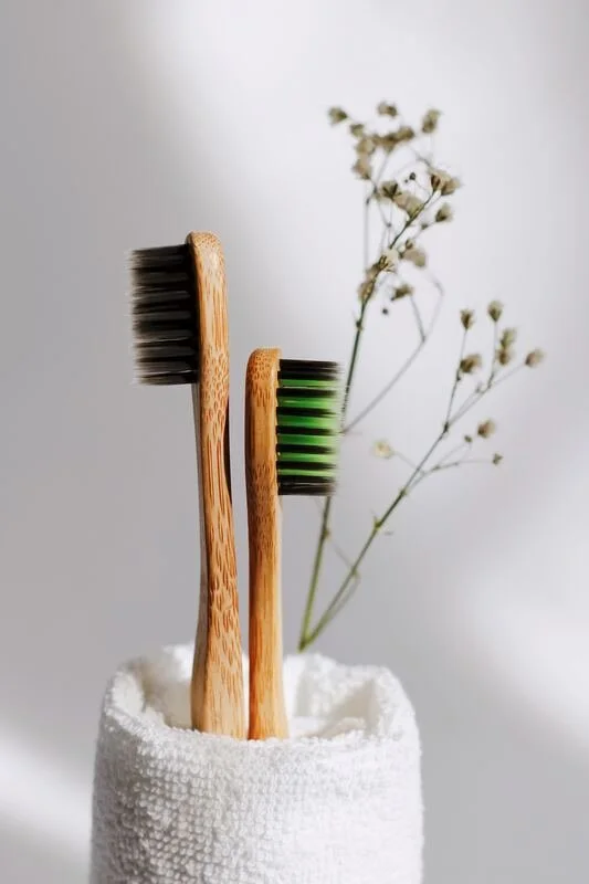 Two bamboo toothbrushes with black and green bristles in a white textured cup, with delicate white flowers in the background.