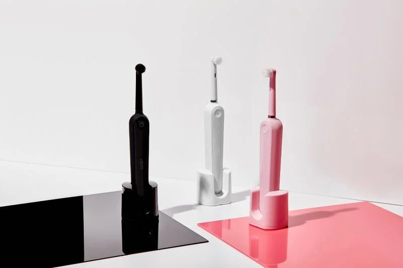 Three electric toothbrushes in black, white, and pink standing on matching charger bases with colored mats underneath in a minimalistic setting.