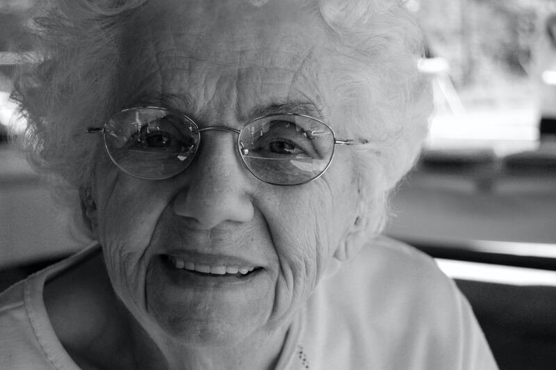 Close-up black and white photo of an elderly woman with curly white hair, wearing round glasses and a light-colored shirt, smiling.