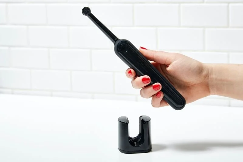 A person's hand holding a black electric toothbrush with an interchangeable brush head, positioned above a charging stand on a white surface against a white tiled wall.