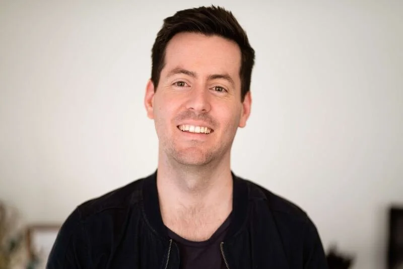 A smiling man with short dark hair, wearing a black jacket, standing indoors with a blurred background.