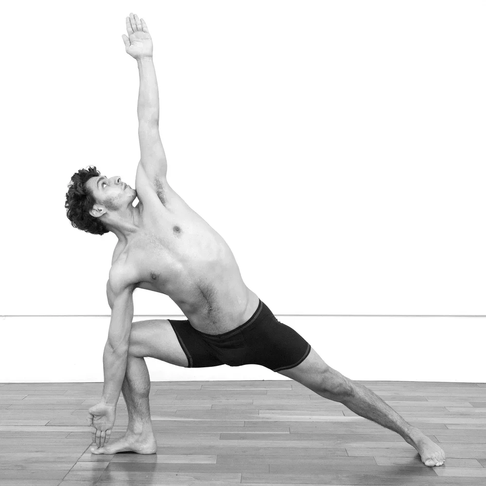 A shirtless man wearing black shorts practicing yoga indoors on a wooden floor, performing a side angle pose with one arm extended upward and the other hand on the floor.
