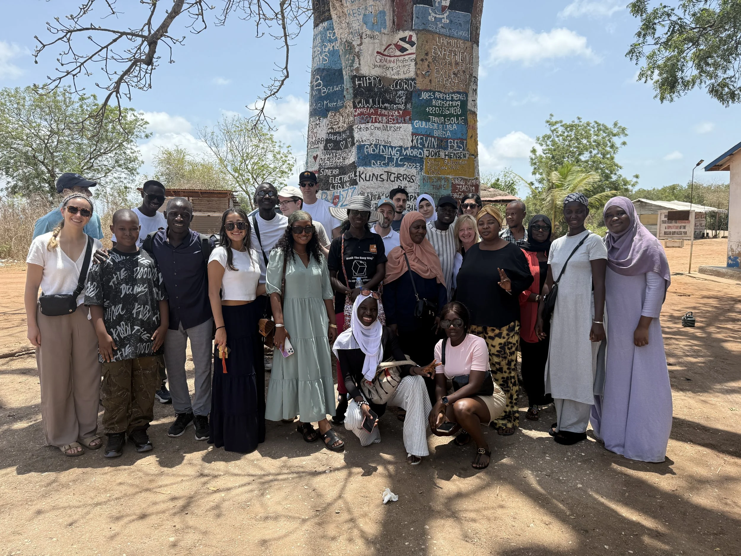 A diverse group of people gathered outdoors under a large tree with graffiti-covered support pole in the background. The group is smiling and posing for a photo on a sunny day in an open area with some small buildings around.