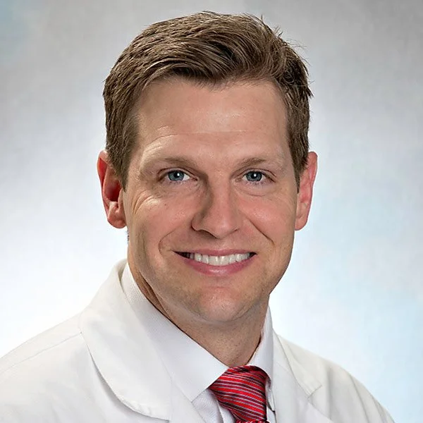 A smiling man in a white lab coat with a red tie, short brown hair and blue eyes, against a neutral background.