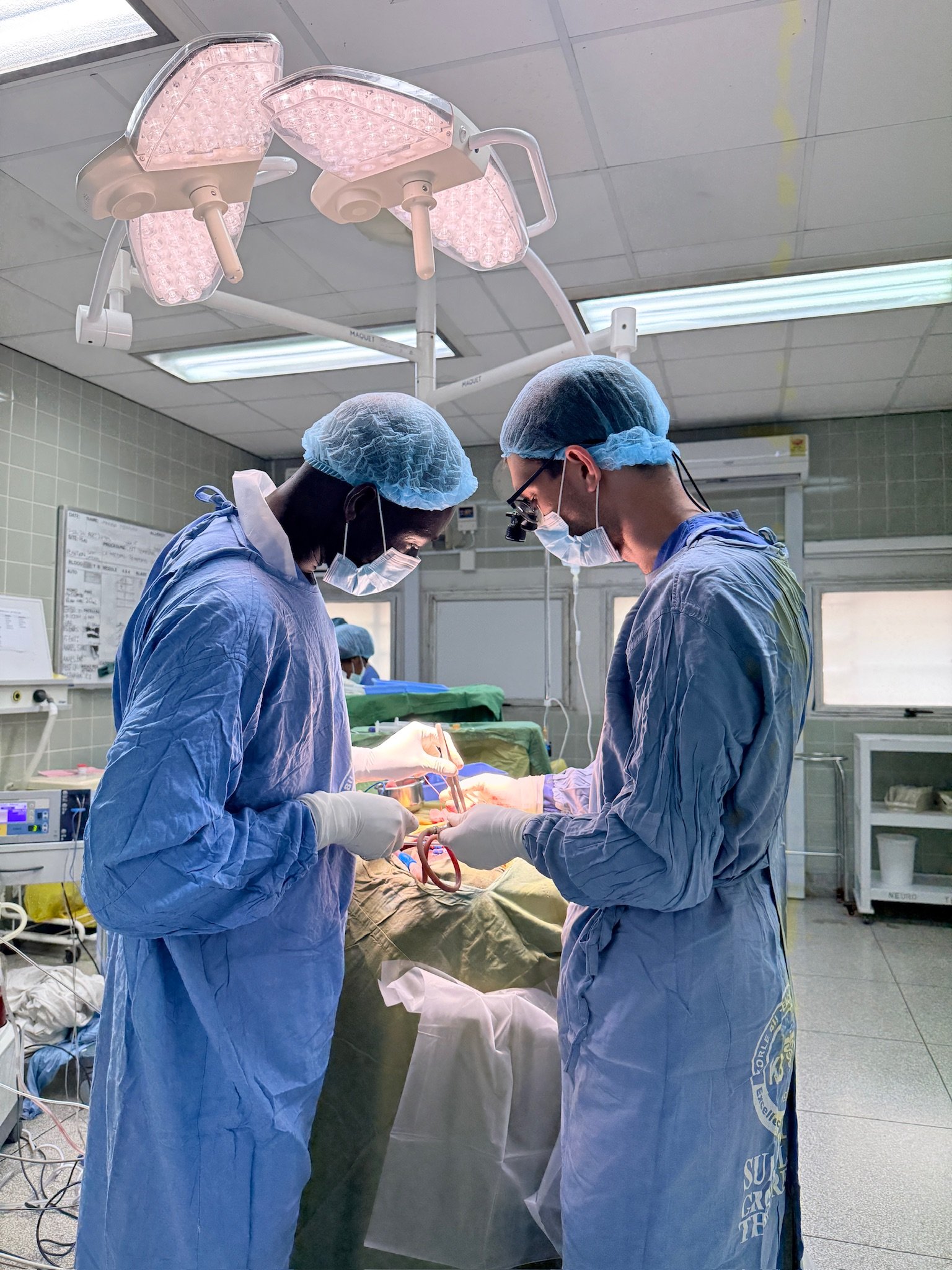 Two surgeons are performing a medical procedure in an operating room, wearing surgical masks, gloves, and gowns, with overhead surgical lights illuminating their work.
