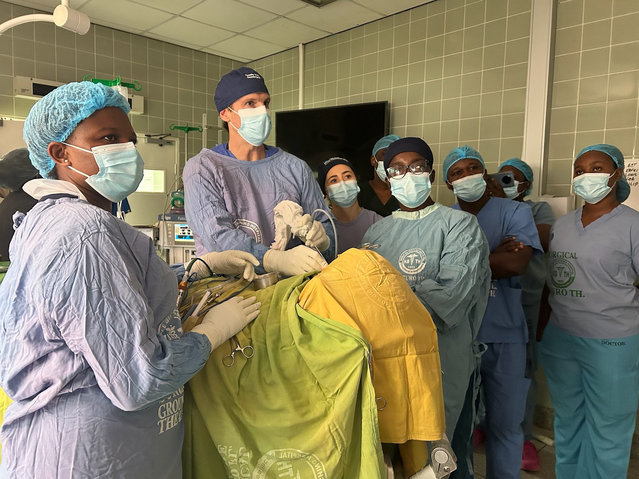 A group of medical professionals in surgical scrubs, masks, and caps gathered around a patient in a hospital operating room.