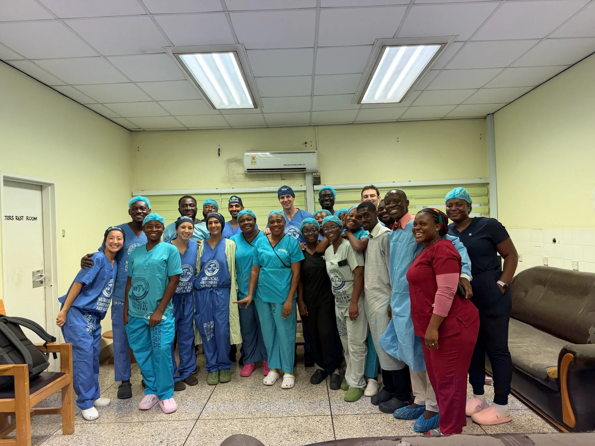 Group of smiling medical professionals and staff in scrubs and masks posing together in a clinic or hospital room.