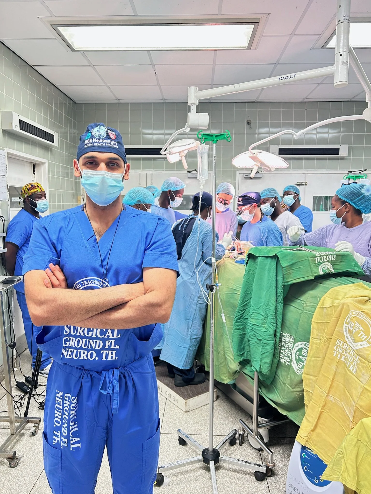 A young male surgeon in blue scrubs and a face mask standing with arms crossed in an operating room, facing camera. Behind him, a team of medical professionals wearing masks and scrubs are gathered around an operating table, preparing for surgery.
