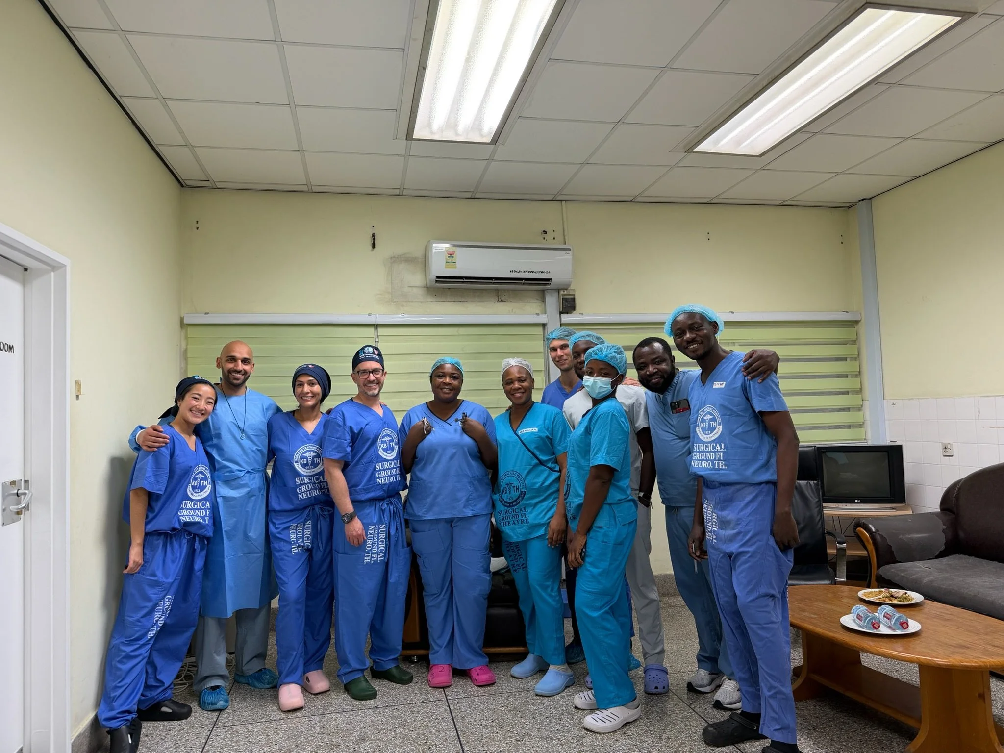 Group of healthcare professionals in scrubs and surgical caps posing together in a room with fluorescent lighting and a window with blinds, some smiling and some wearing face masks.