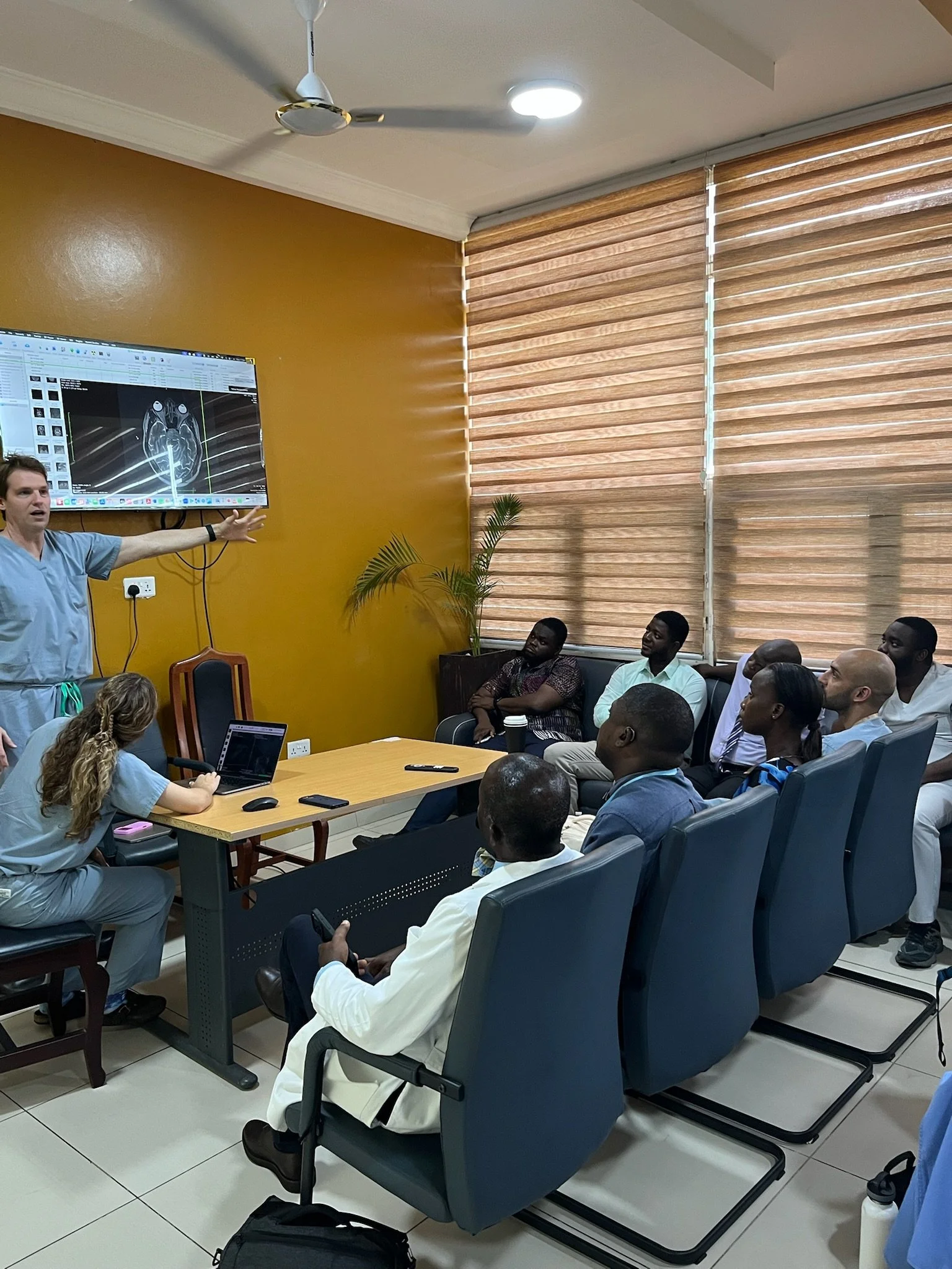 A group of people attending a presentation or lecture in a conference room. The presenter, wearing medical scrubs, is pointing at a screen displaying medical imaging.