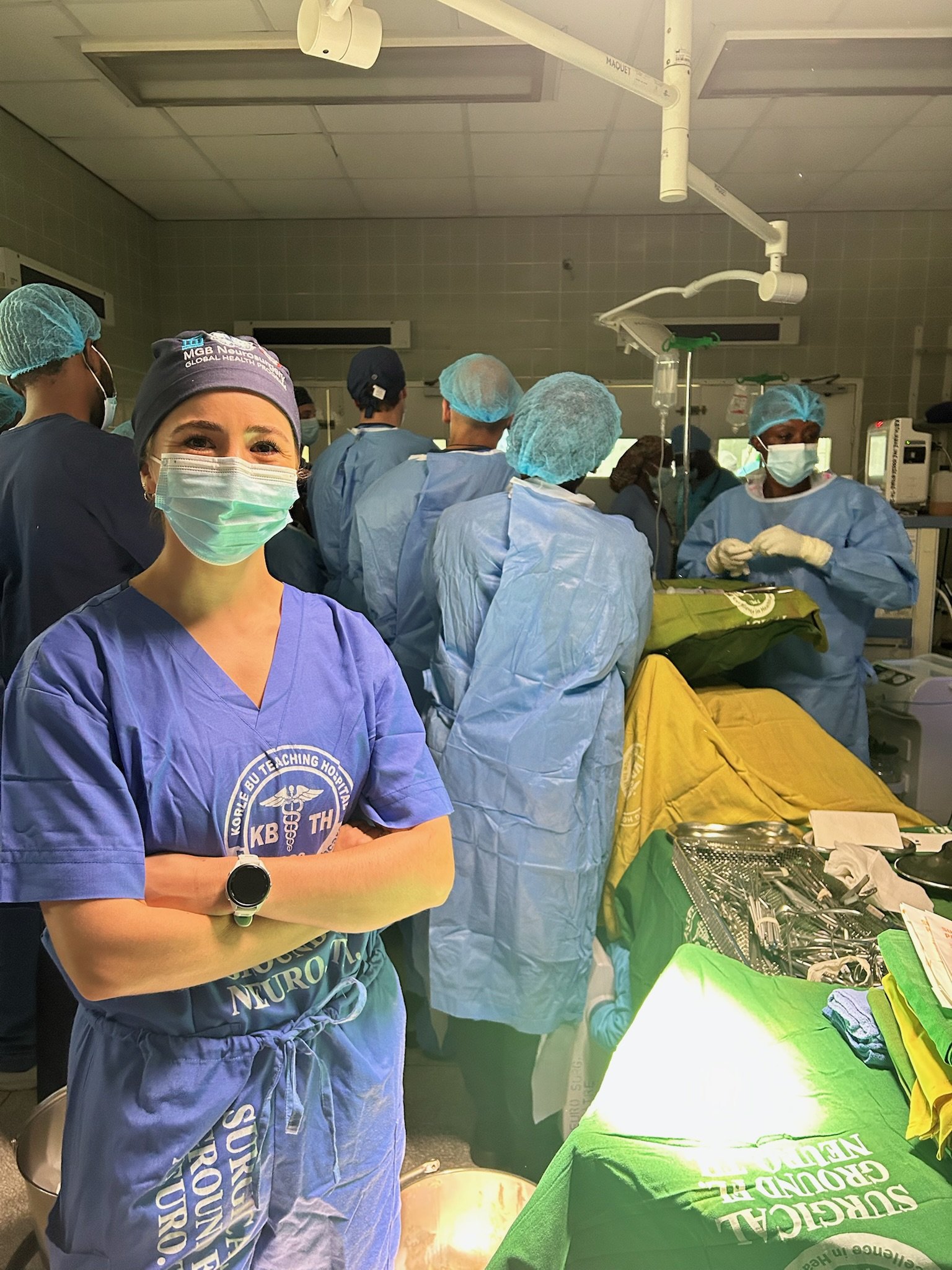 Healthcare workers wearing scrubs, masks, and hair covers in a medical setting, preparing for surgery or medical procedure with surgical tools and equipment on a green covered table.